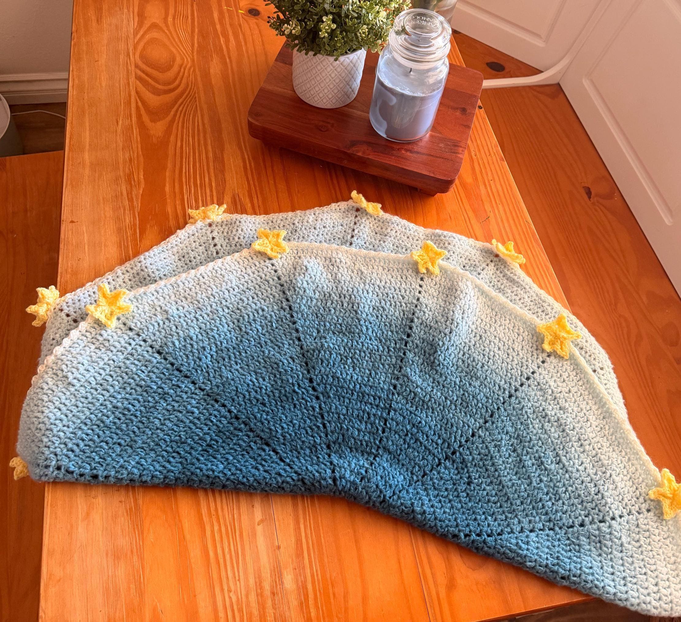 A light blue and white crochet blanket with yellow star appliques is folded in half on a wooden table.