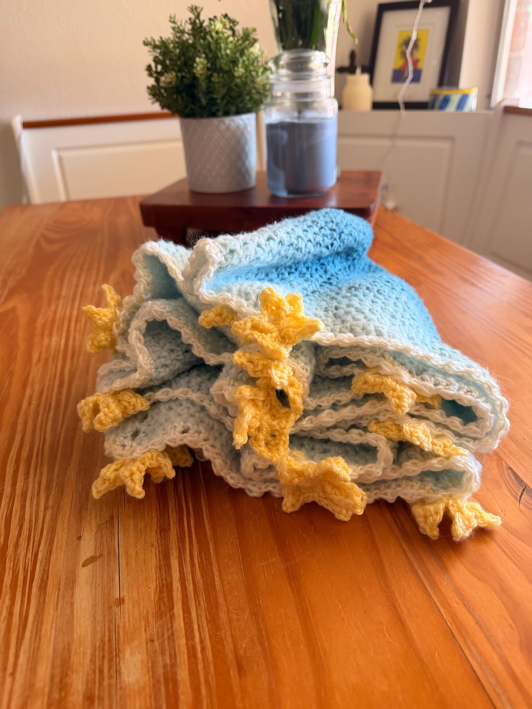 A light blue and white crochet blanket with yellow star appliques is folded into a stack on a wooden table.