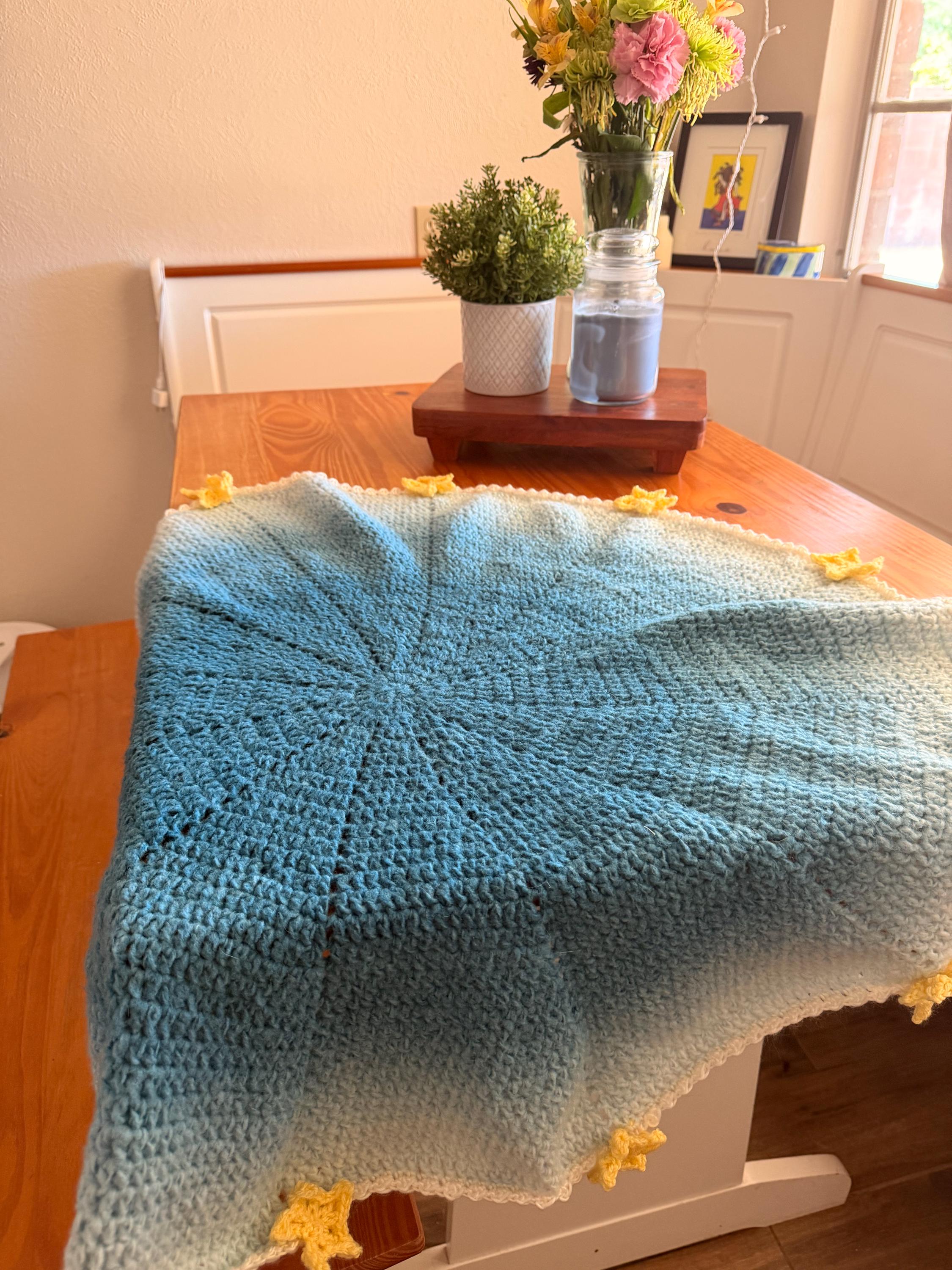 A light blue and white crochet blanket with yellow star appliques is spread out on a wooden table, showing its circular center.