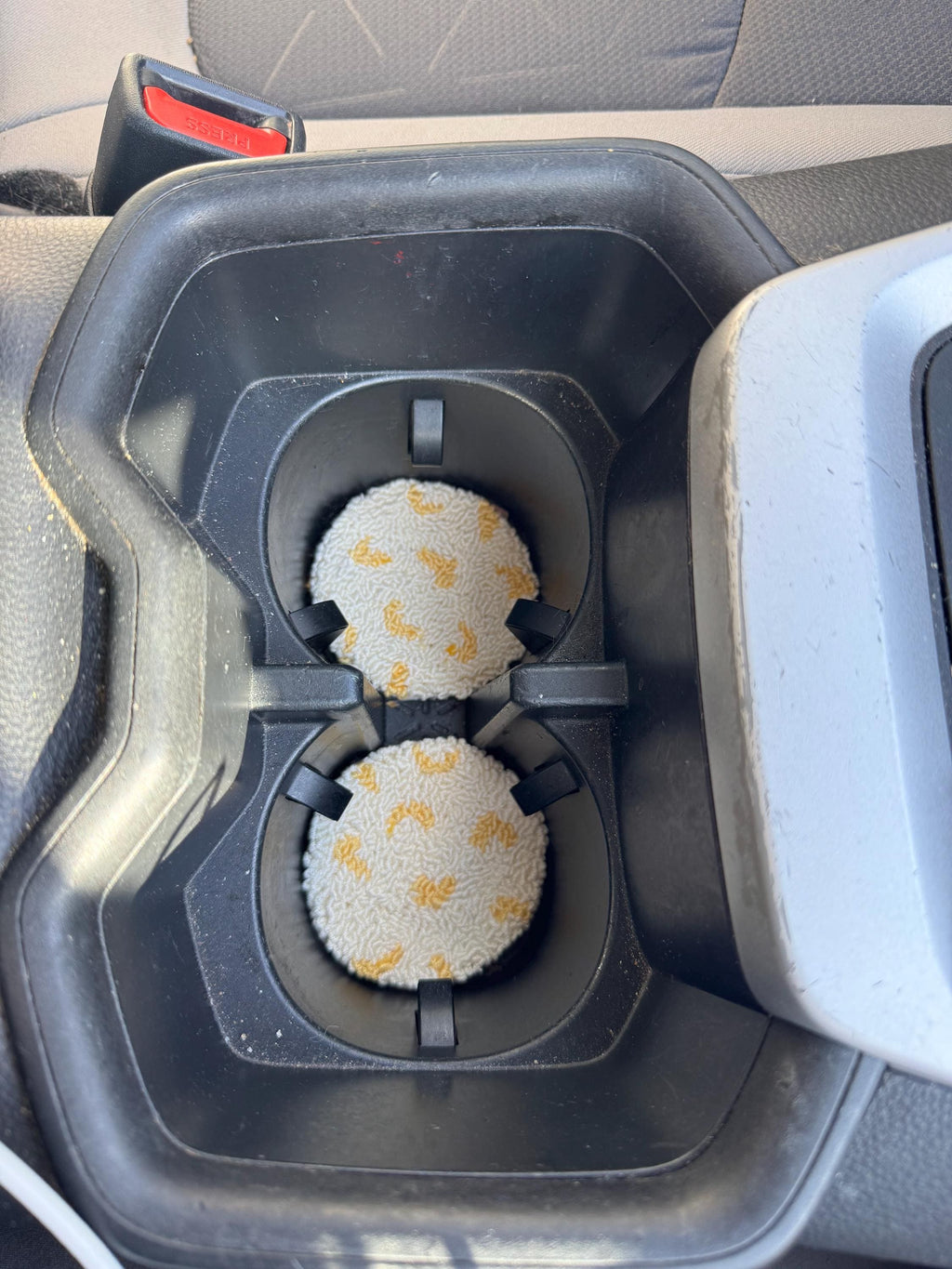 Two round punch needle coasters with a yellow fruit pattern are placed in the cup holders of a car's center console.