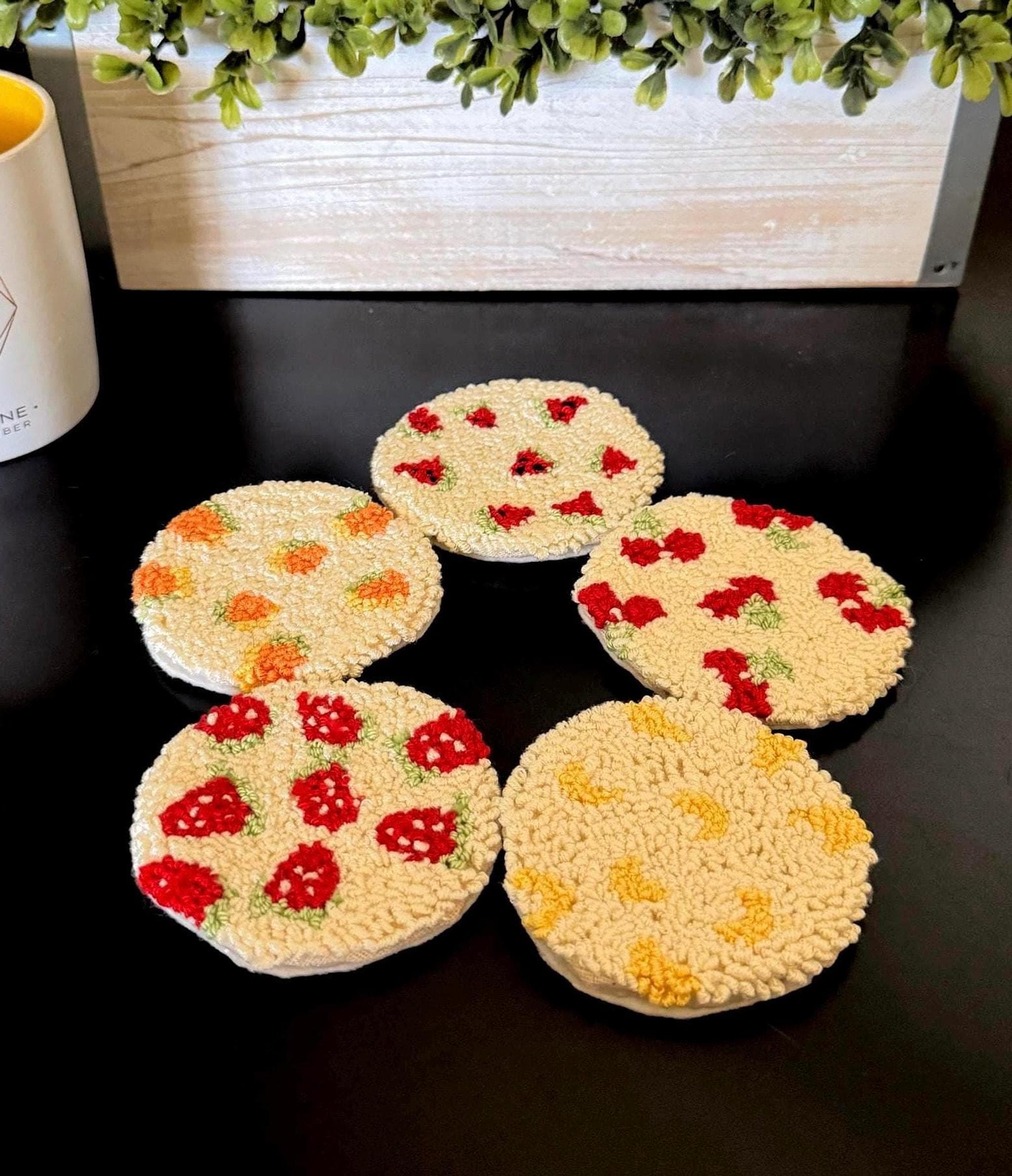 Five round punch needle coasters with fruit designs, including strawberries, oranges, and yellow fruit, are arranged on a black surface.