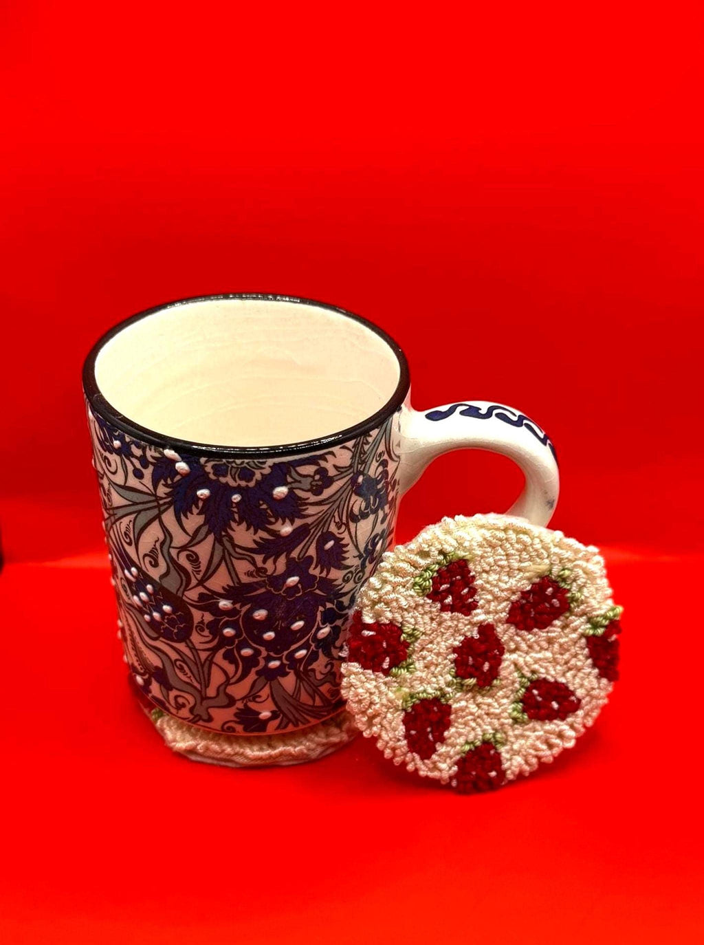 A white and blue patterned ceramic mug sits next to a round punch needle coaster with a red strawberry design on a red background.