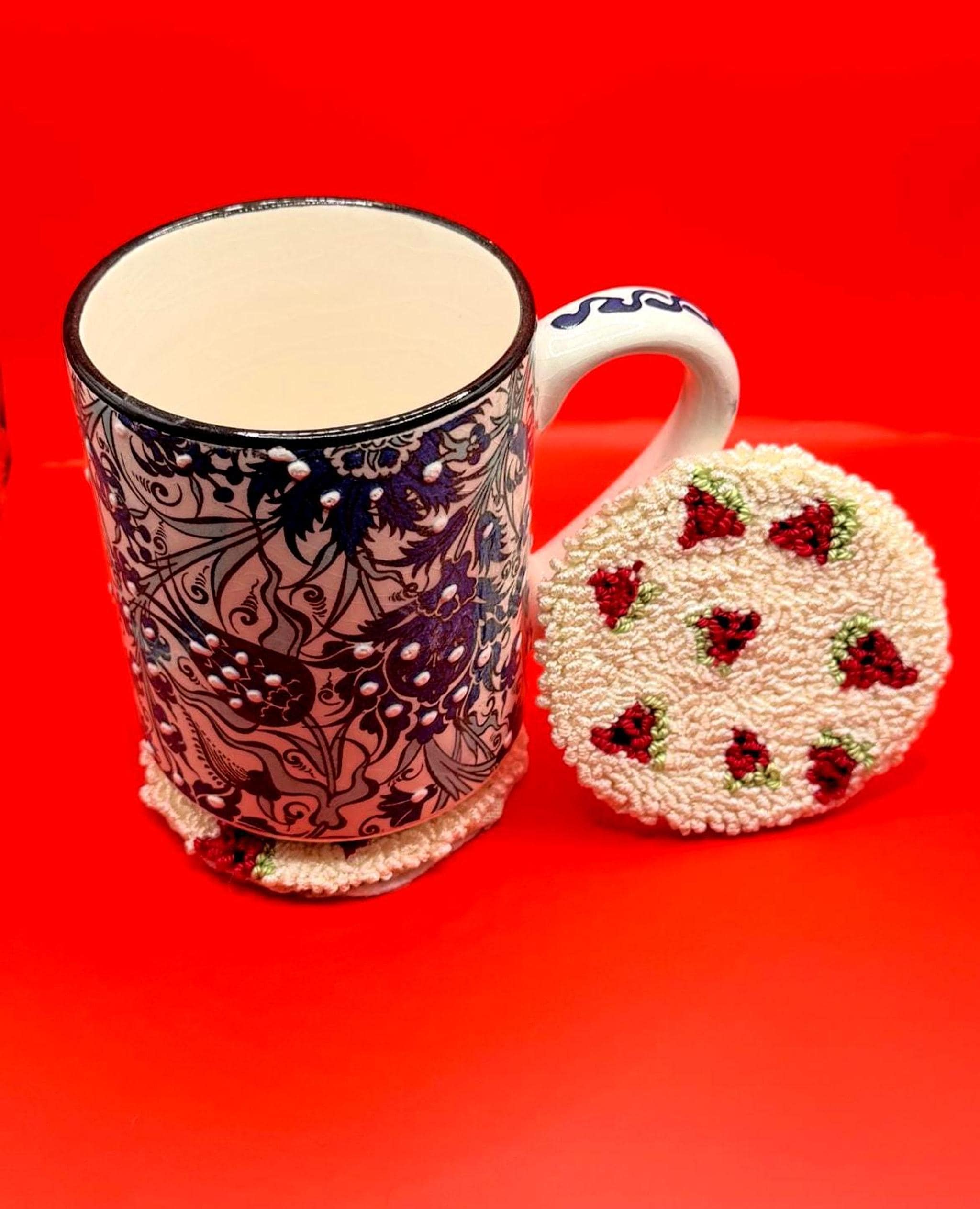 A white and blue patterned ceramic mug is next to a round punch needle coaster with a red strawberry design on a red background.