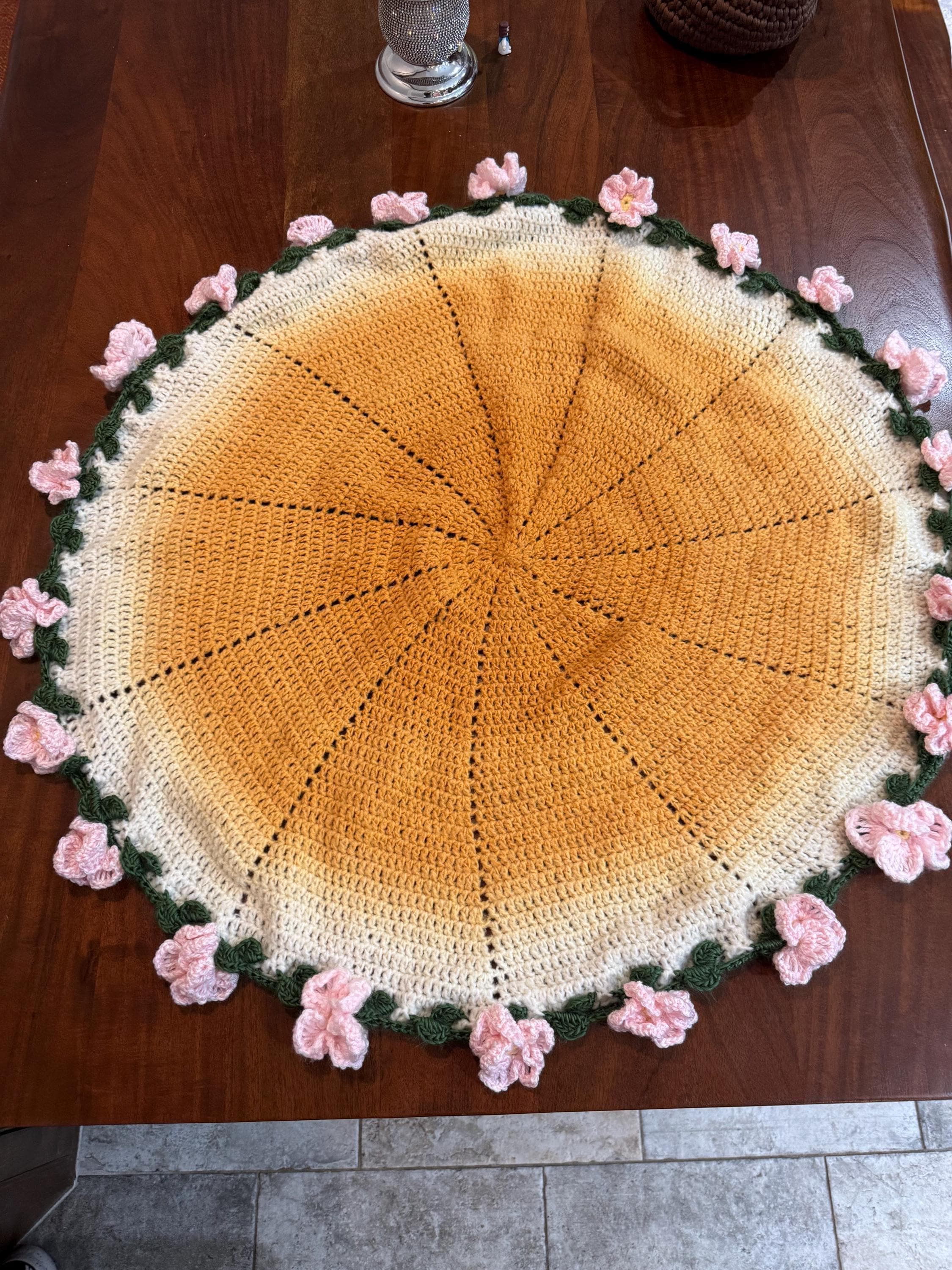 A round, yellow and white crochet blanket with a border of green crochet leaves and pink crochet flowers, on a wooden table.