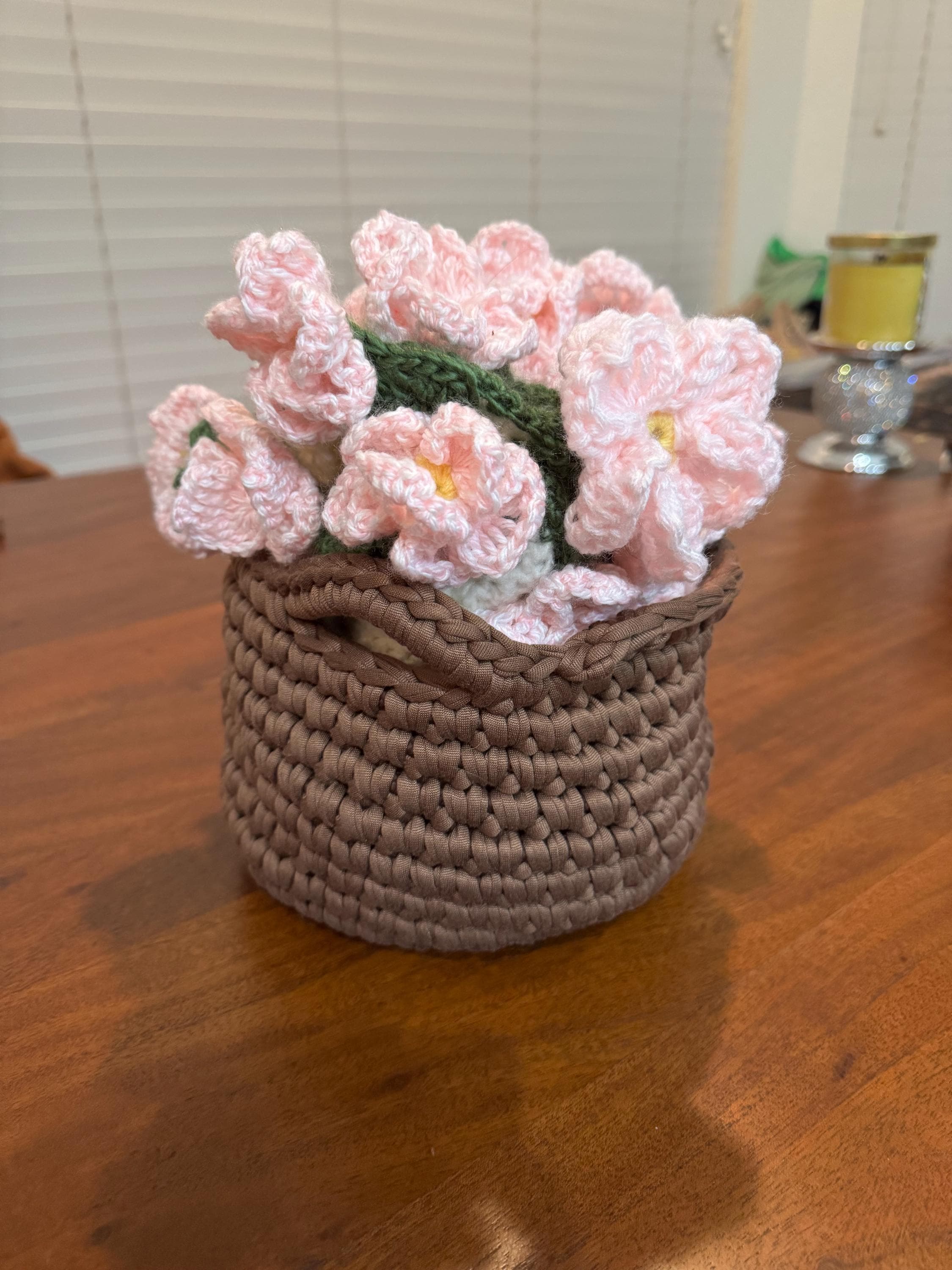 A small brown crochet basket holds a bouquet of pink crochet flowers with green stems, on a wooden table.
