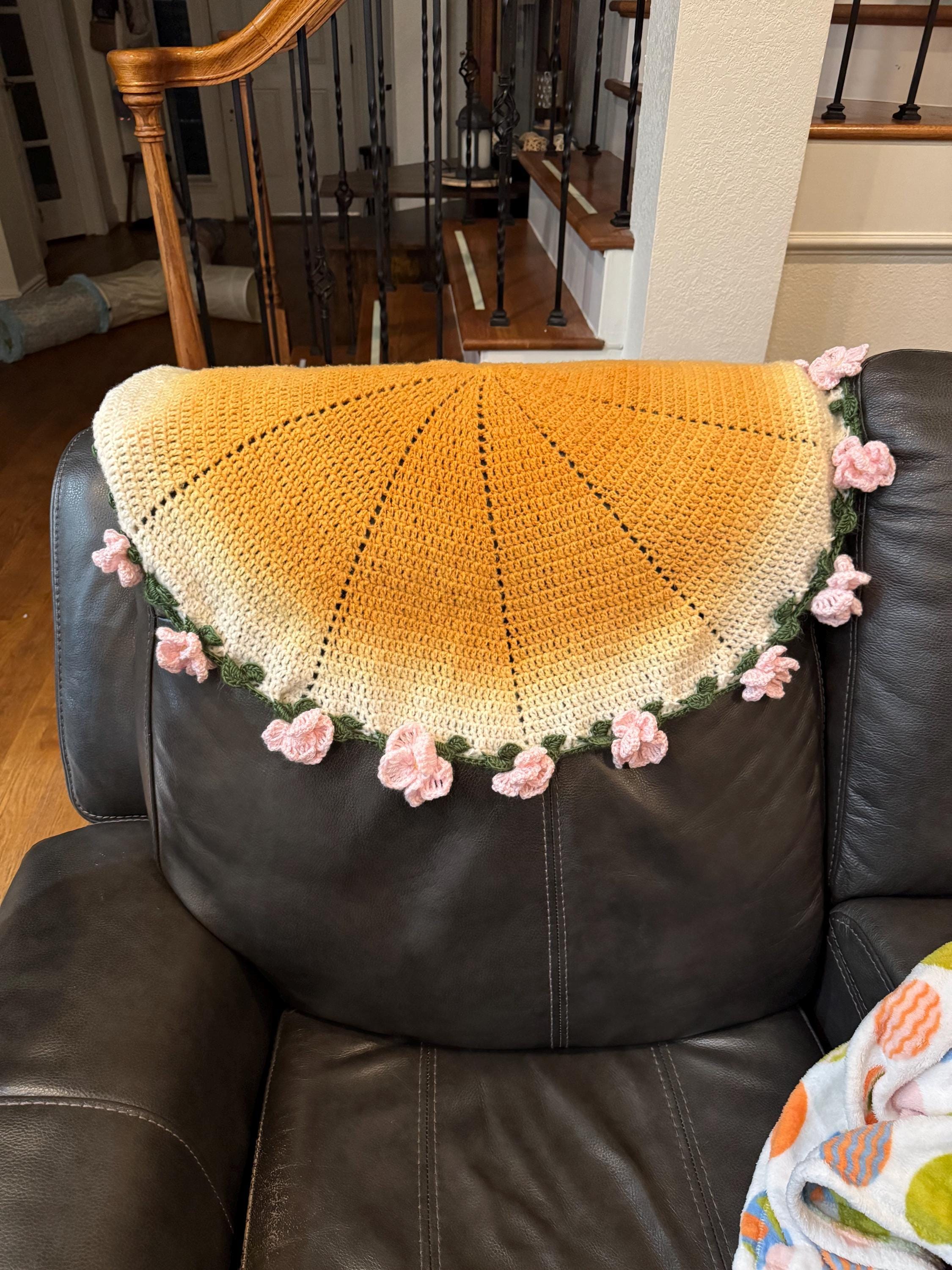 A round, yellow and white crochet blanket with pink crochet flowers along the edge is draped over a dark leather couch.