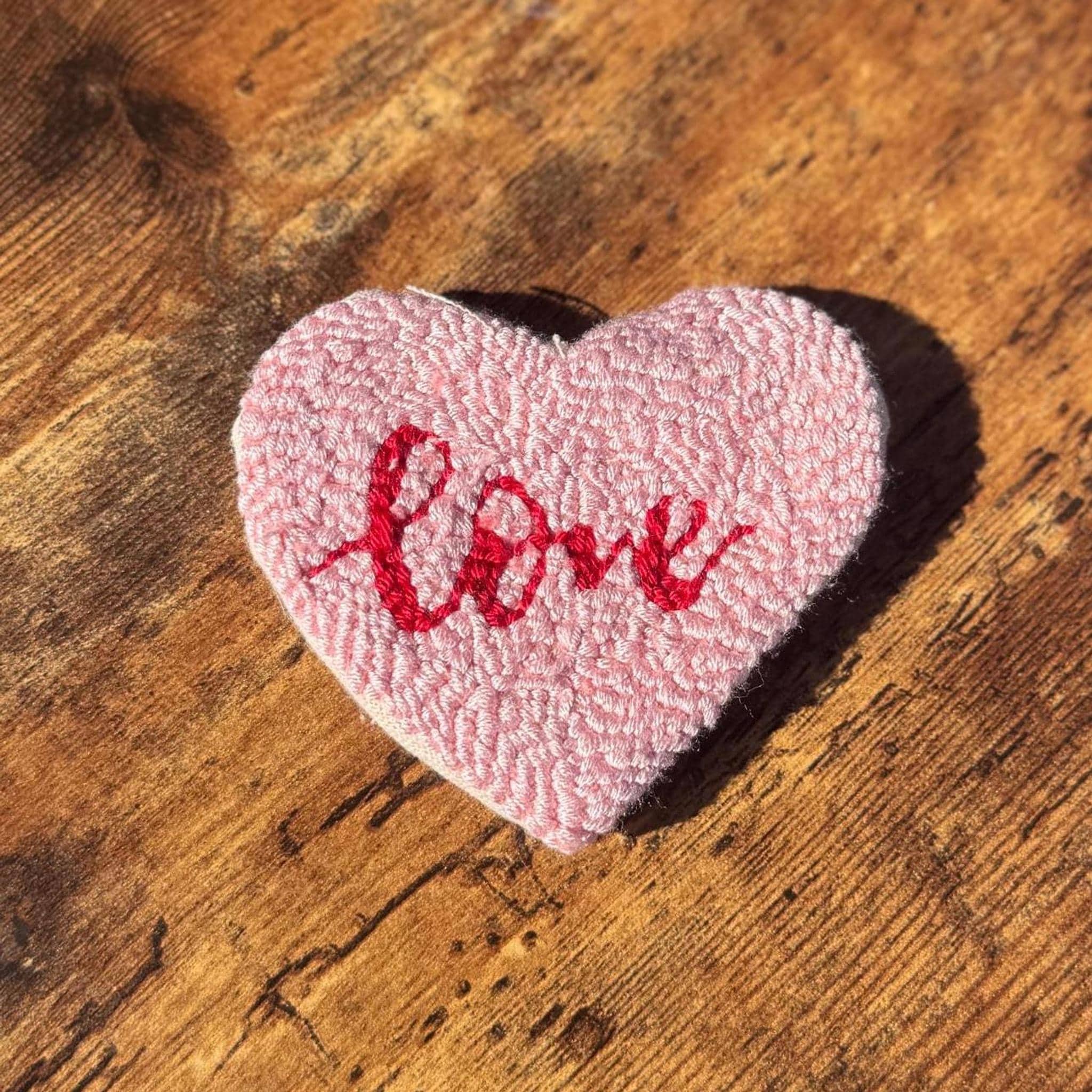 A pink heart-shaped punch needle coaster with the word "love" embroidered in red script on a wooden surface.