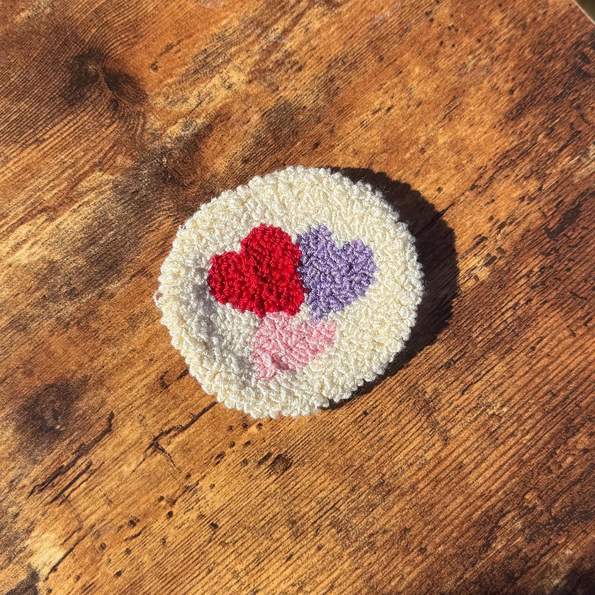 A round white punch needle coaster with three hearts in red, purple, and pink on a wooden surface.