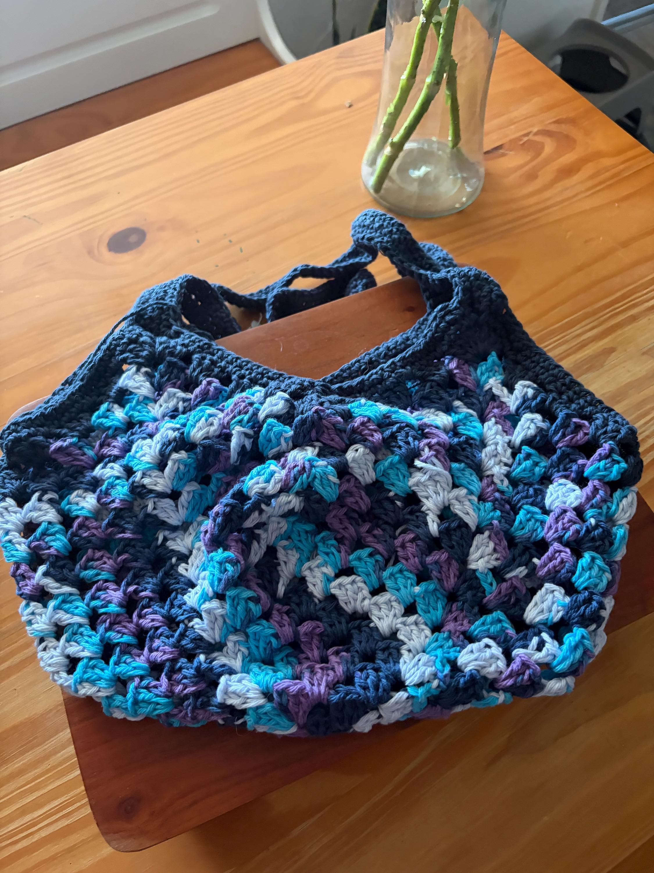 A dark blue crochet market bag with light blue, purple, and white accents is centered on a wooden table.
