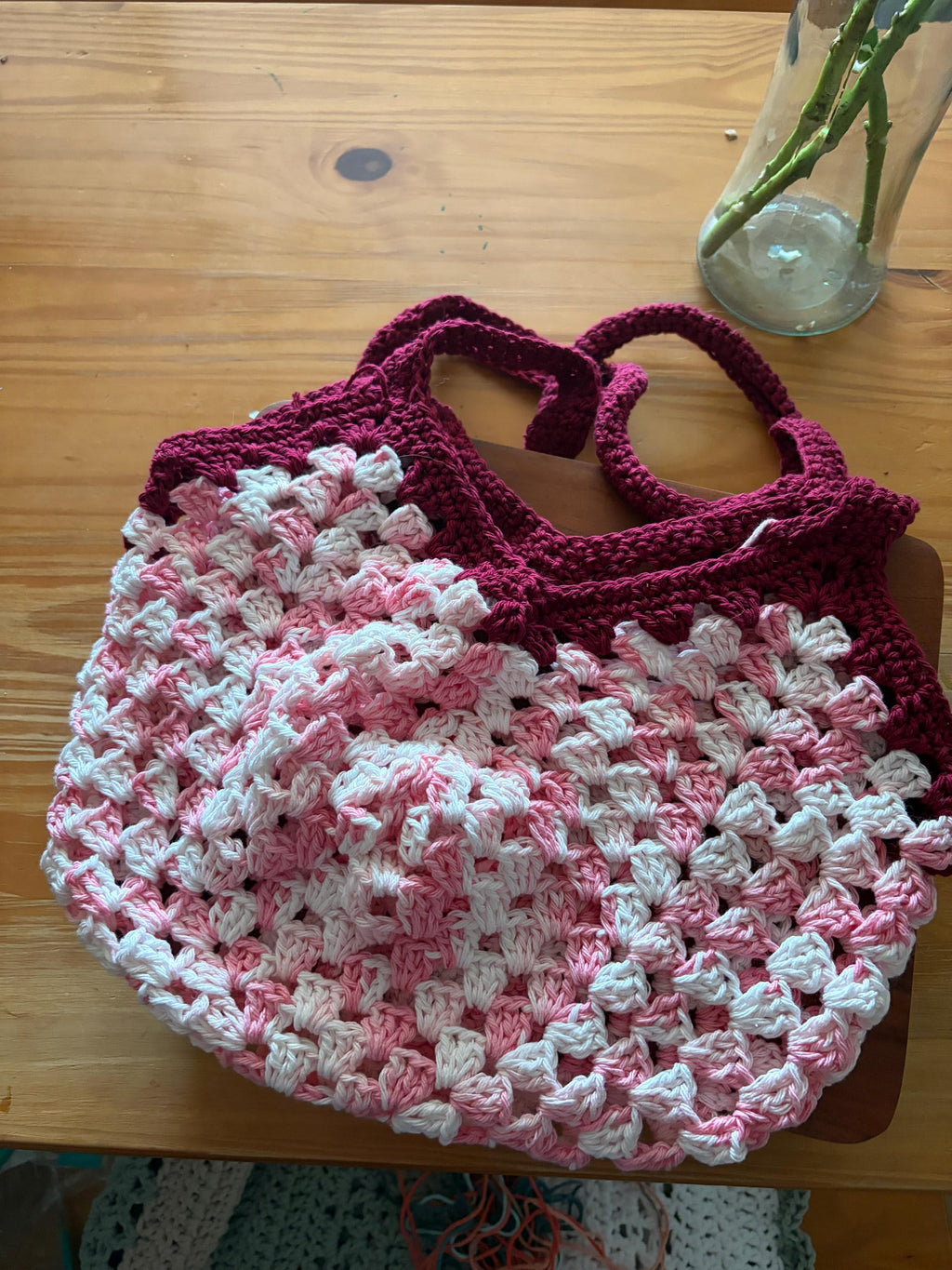 A dark pink crochet market bag with light pink and white accents is centered on a wooden table.