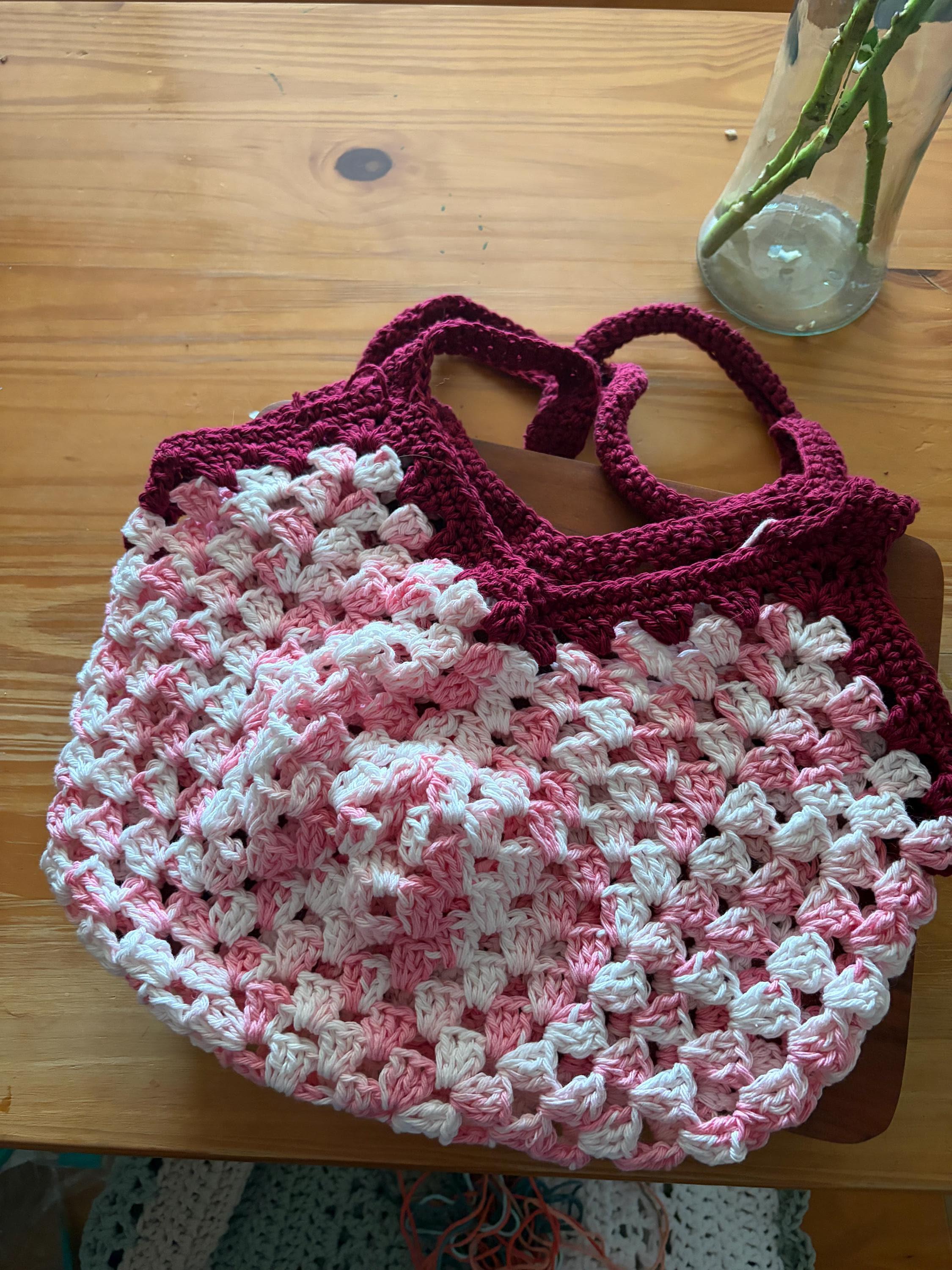 A dark pink crochet market bag with light pink and white accents is centered on a wooden table.