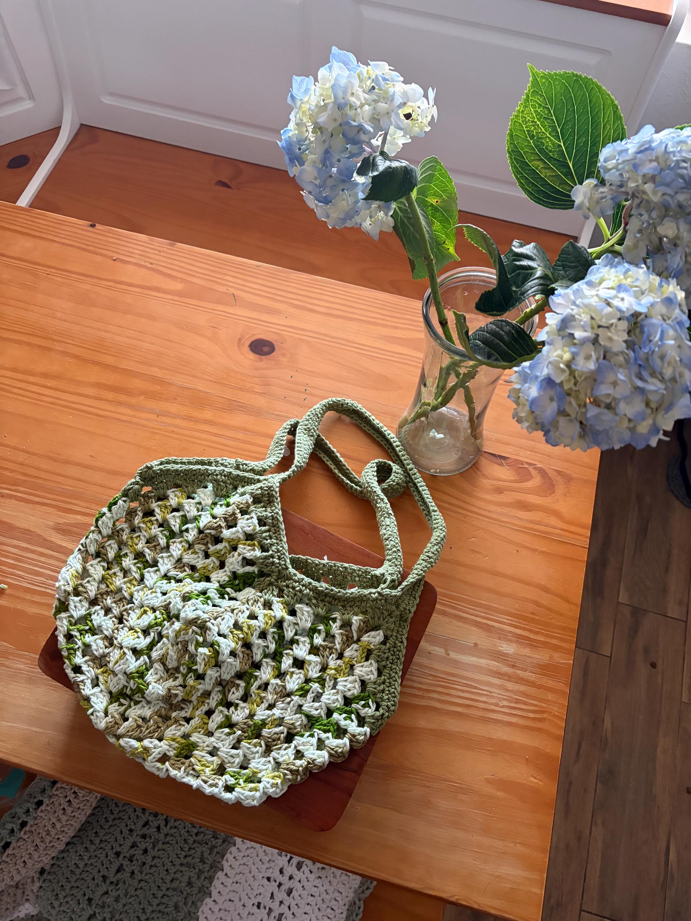A green crochet market bag with white and light green accents sits on a wooden table next to a vase of blue hydrangeas.
