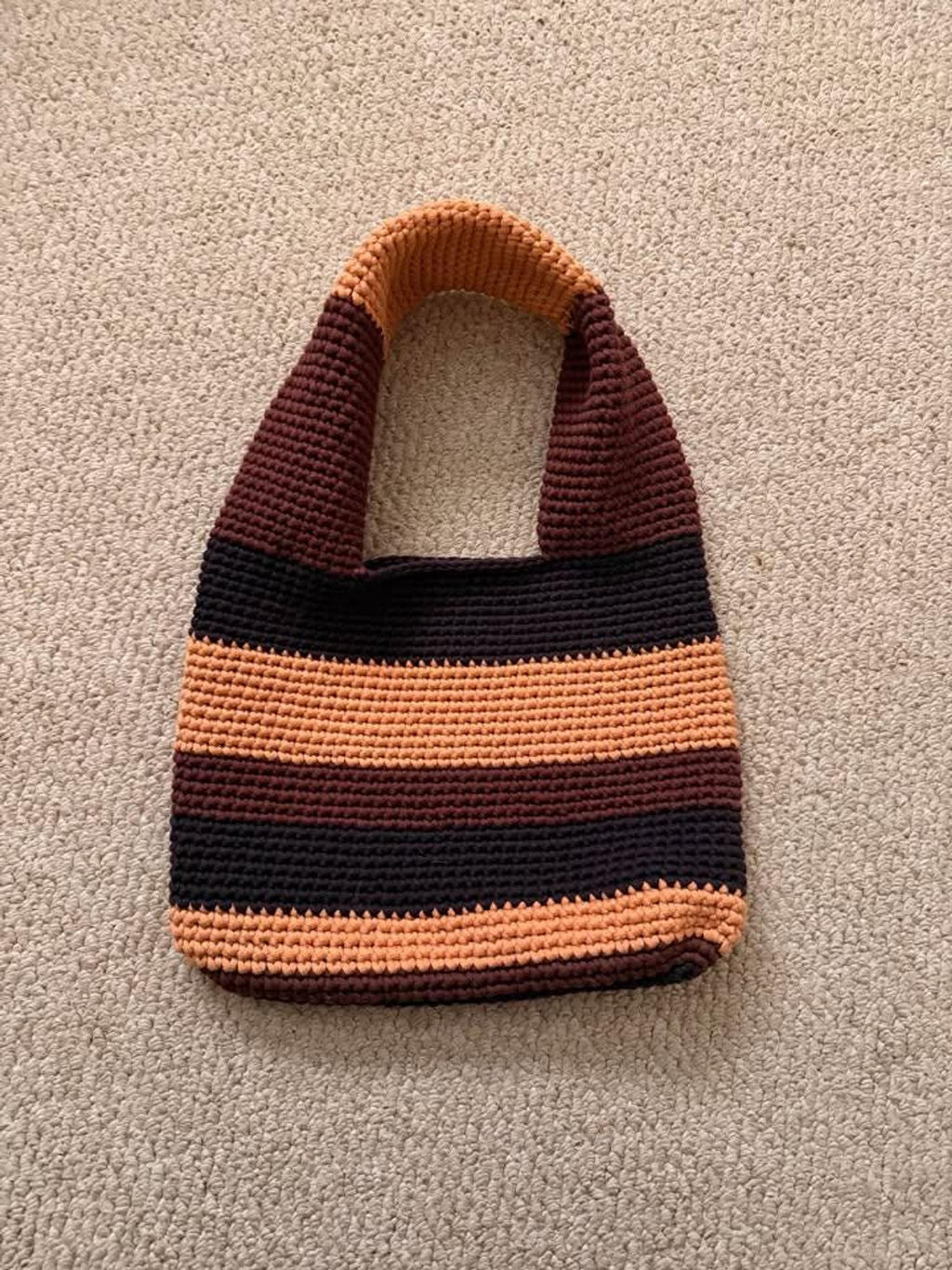 A crocheted tote bag with horizontal stripes in dark brown, light brown, and orange lies flat on a light brown carpet.