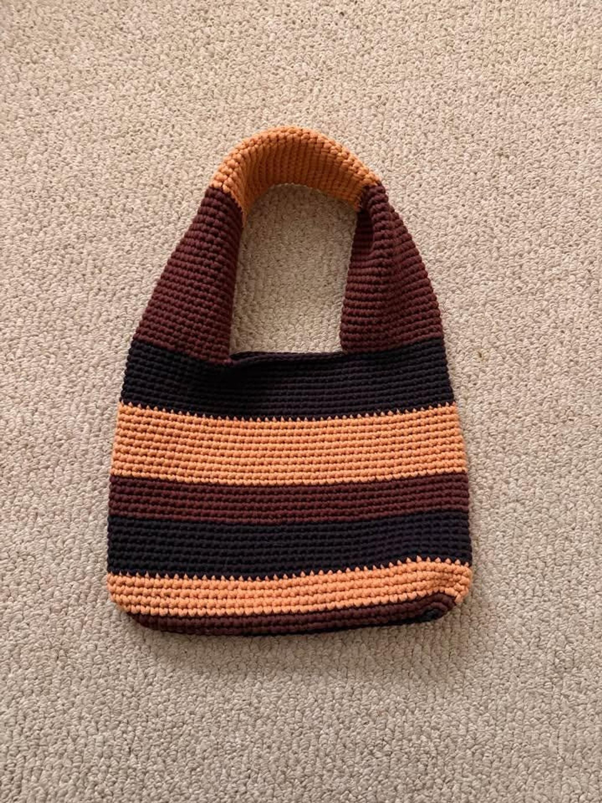 A crocheted tote bag with horizontal stripes in dark brown, light brown, and orange lies flat on a light brown carpet.