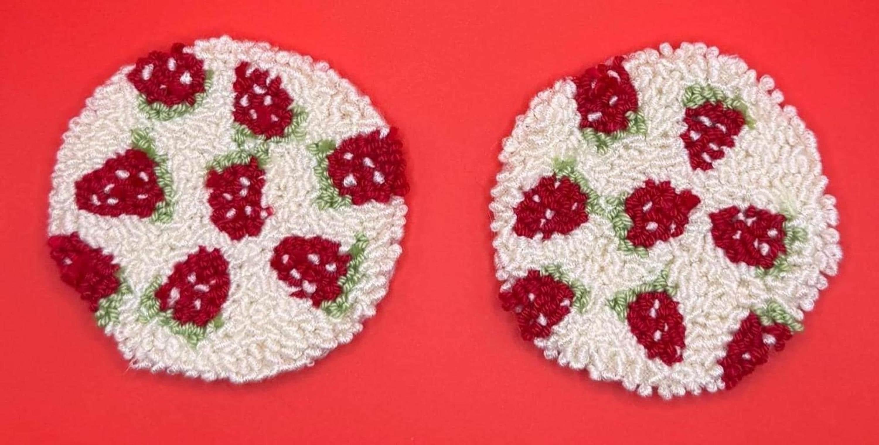 Two round punch needle coasters, each featuring a red strawberry pattern on a cream background, are displayed side-by-side on a red surface.