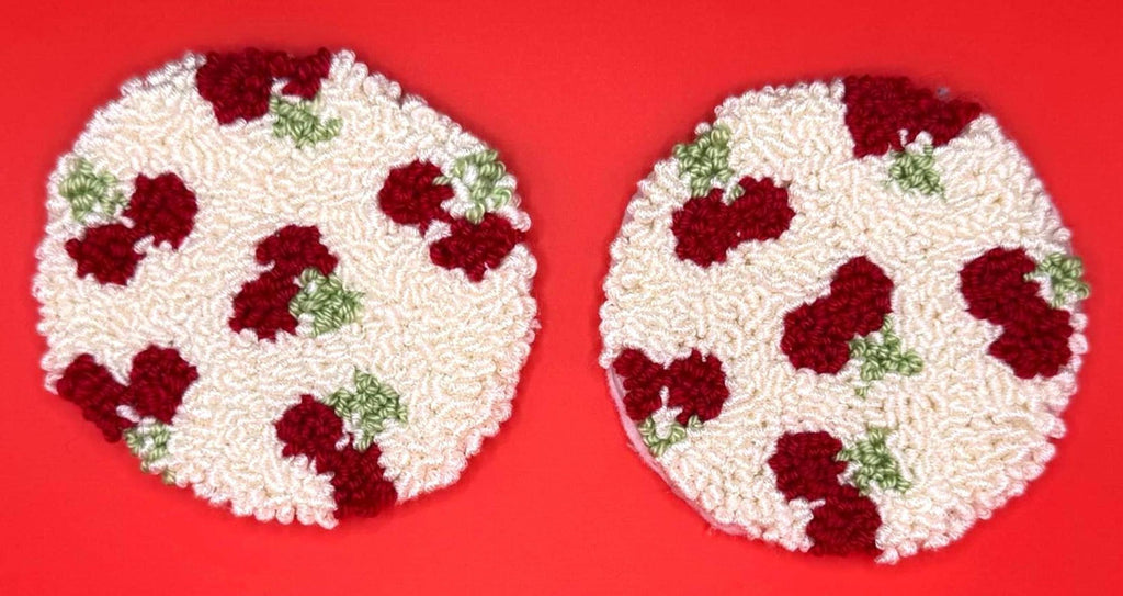 Two round punch needle coasters, each with a red cherry-like fruit pattern on a cream background, are displayed on a red surface.