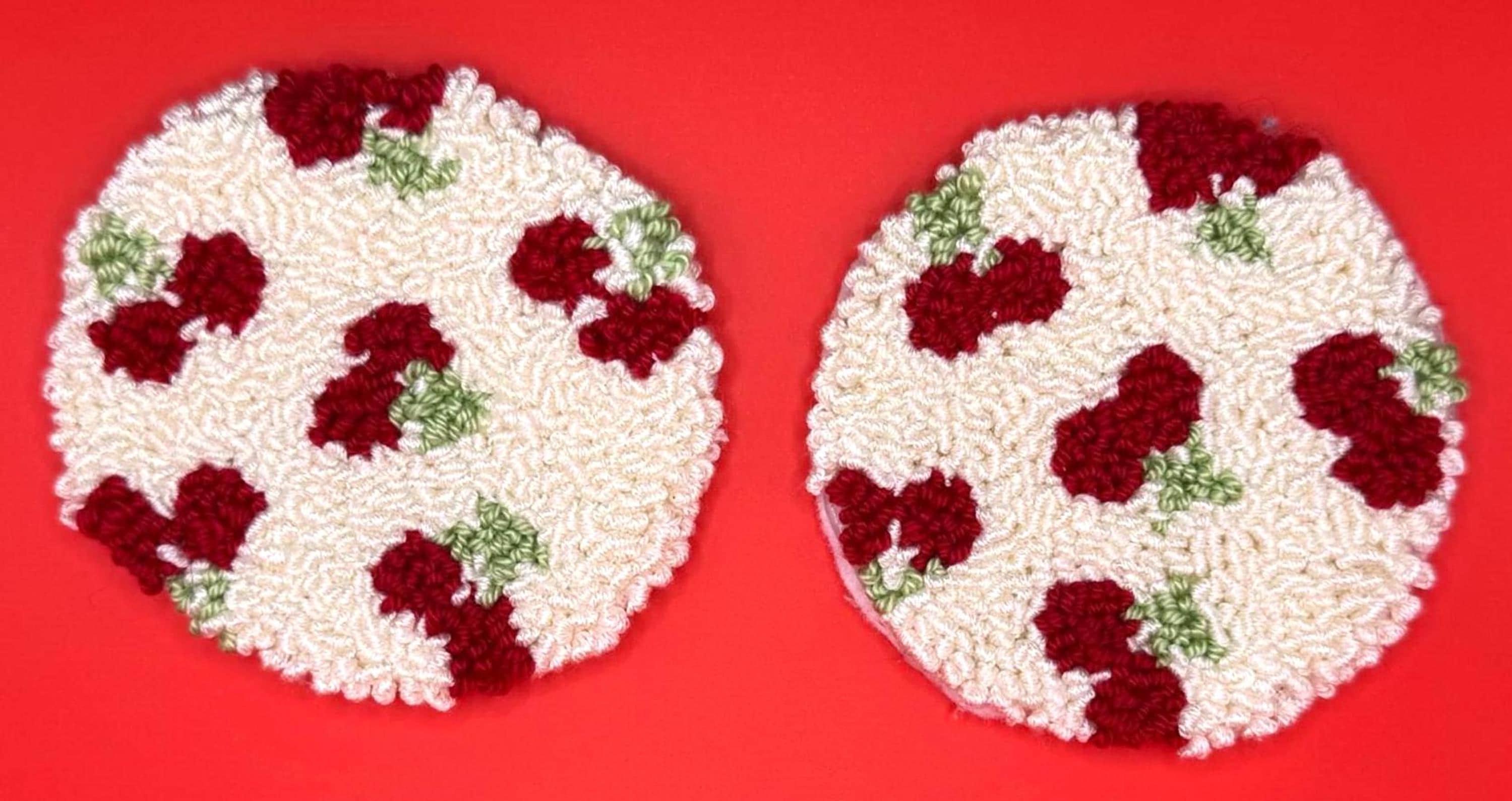 Two round punch needle coasters, each with a red cherry-like fruit pattern on a cream background, are displayed on a red surface.