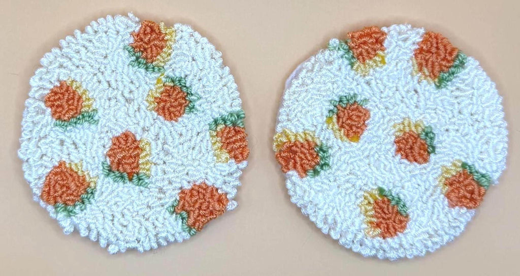 Two round punch needle coasters, each with an orange fruit pattern on a cream background, are displayed side-by-side.