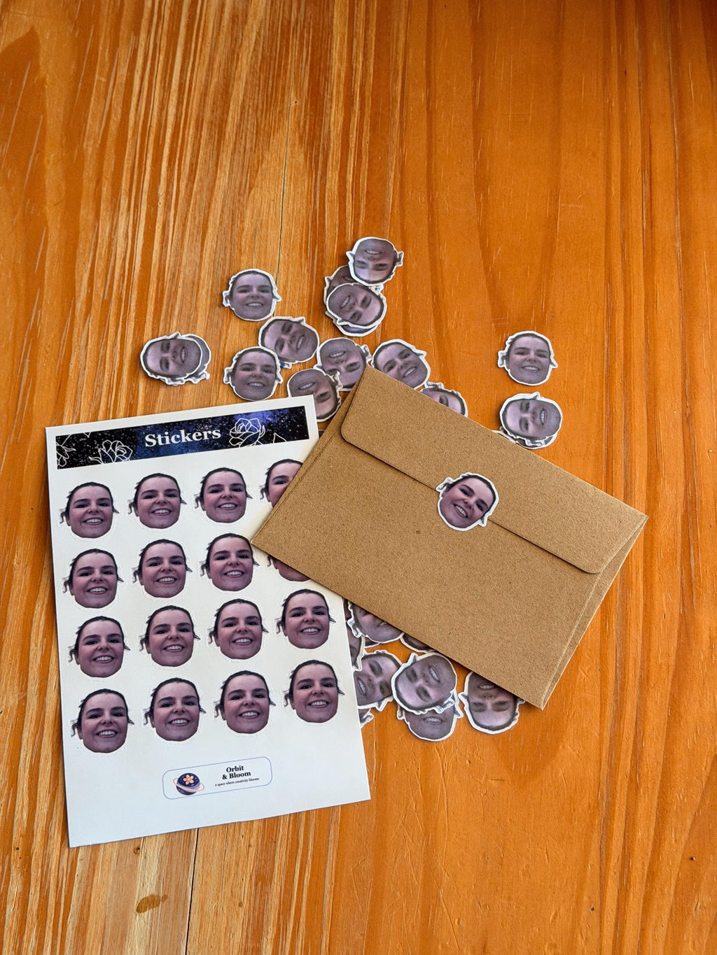 A sheet of custom face stickers, a brown paper envelope sealed with one of the face stickers, and numerous loose face confetti pieces scattered on a wooden table.