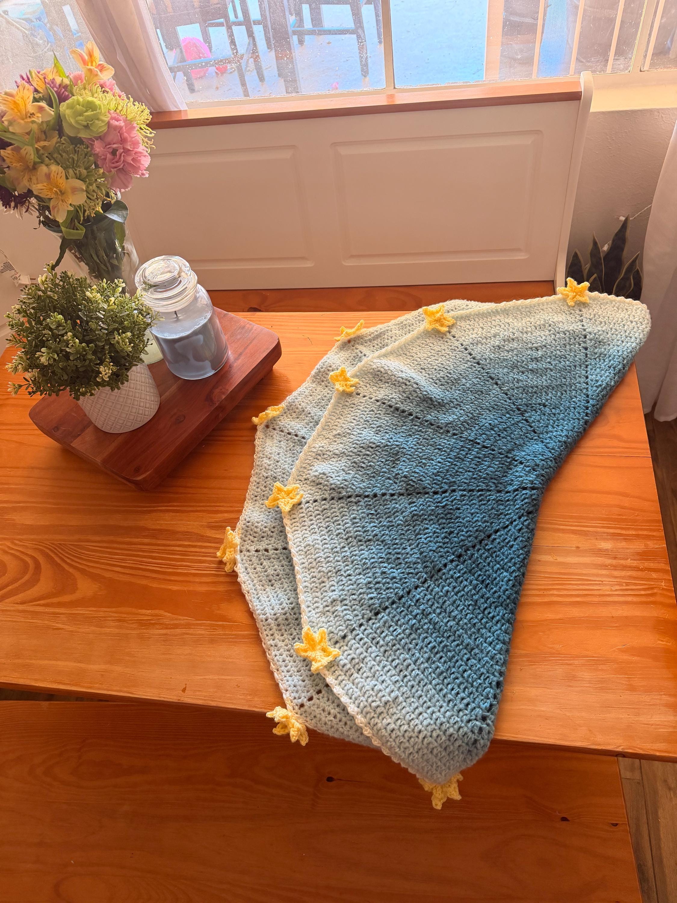 A light blue and white crochet blanket with yellow star appliques is draped over a wooden table, with one side folded over.