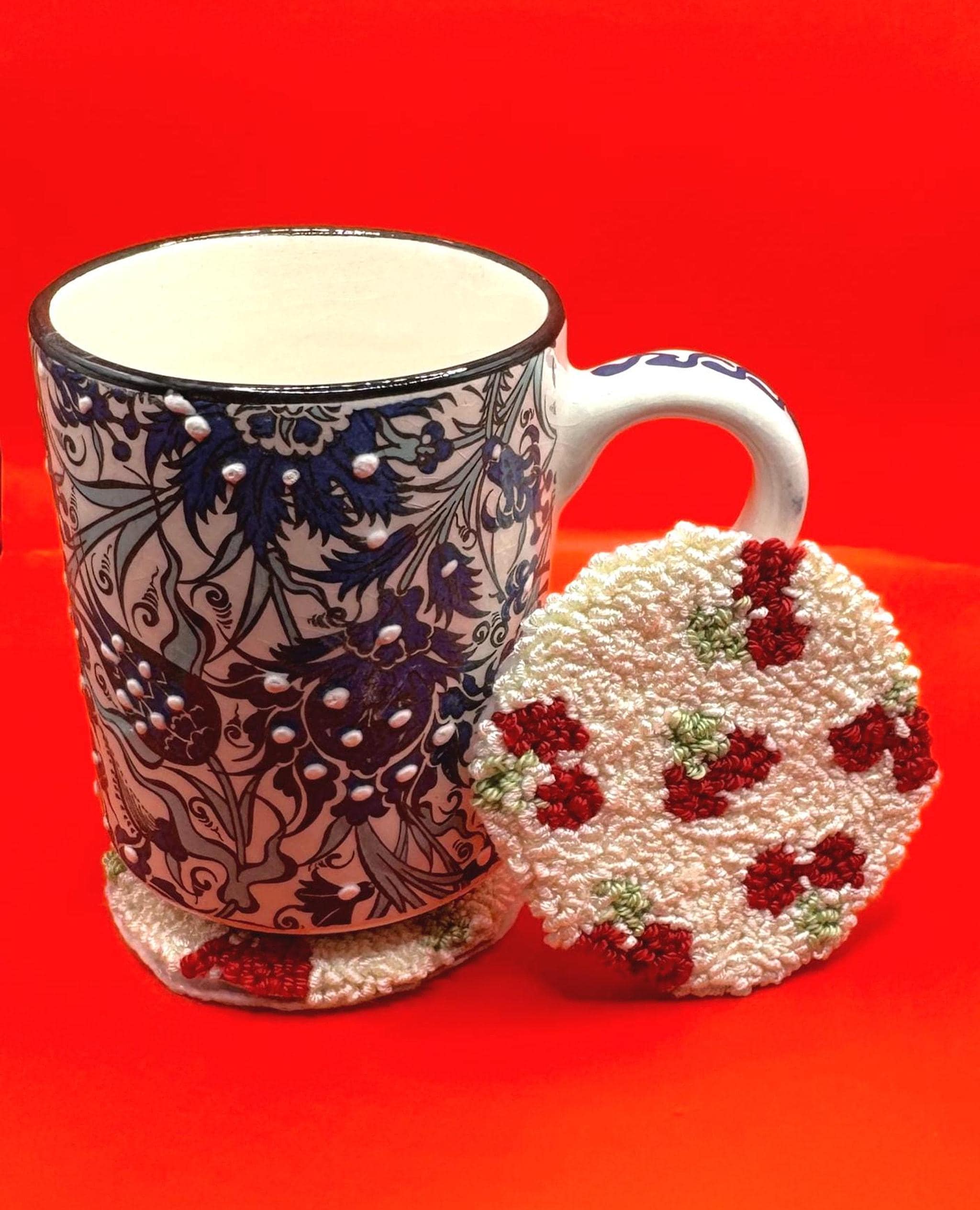A white and blue patterned ceramic mug is next to a round punch needle coaster with a red strawberry design on a red background.