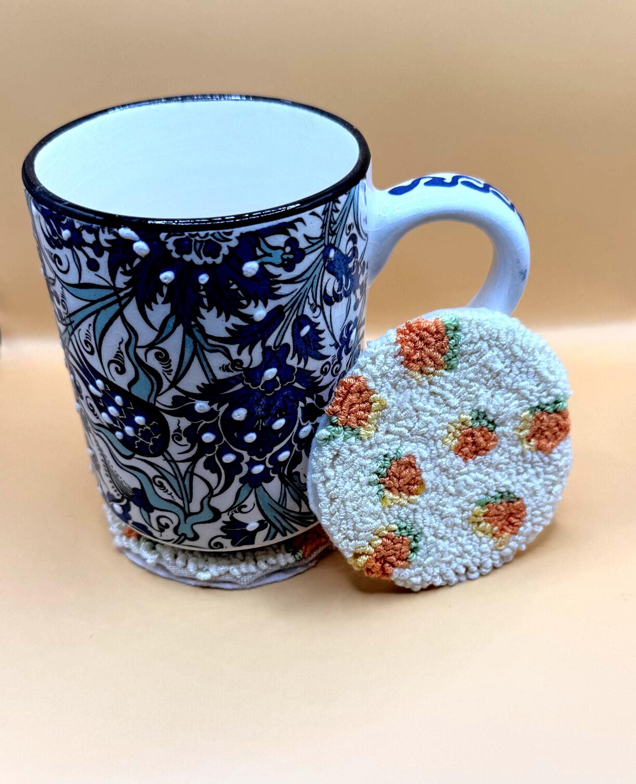 A white and blue patterned ceramic mug sits next to a round punch needle coaster with an orange fruit design on a cream background.