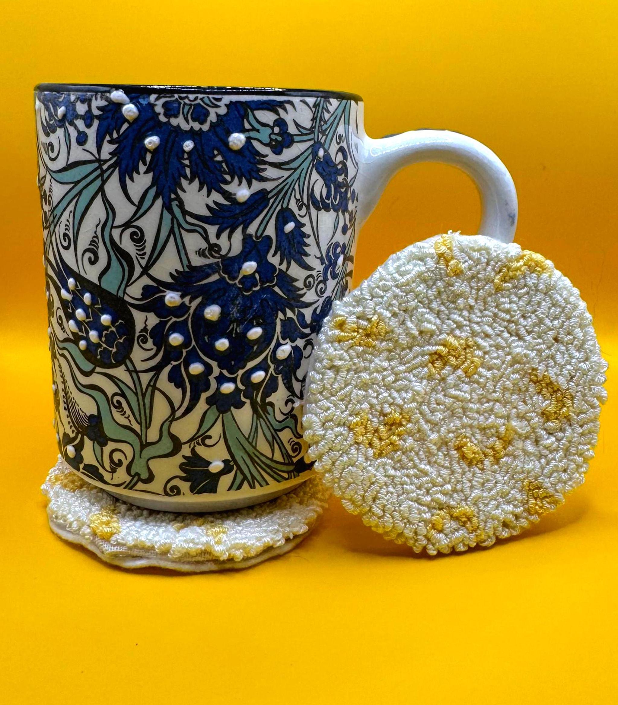 A white mug with blue floral patterns sits on a round, white and yellow punch needle coaster, with another matching coaster next to it, all on a yellow background.