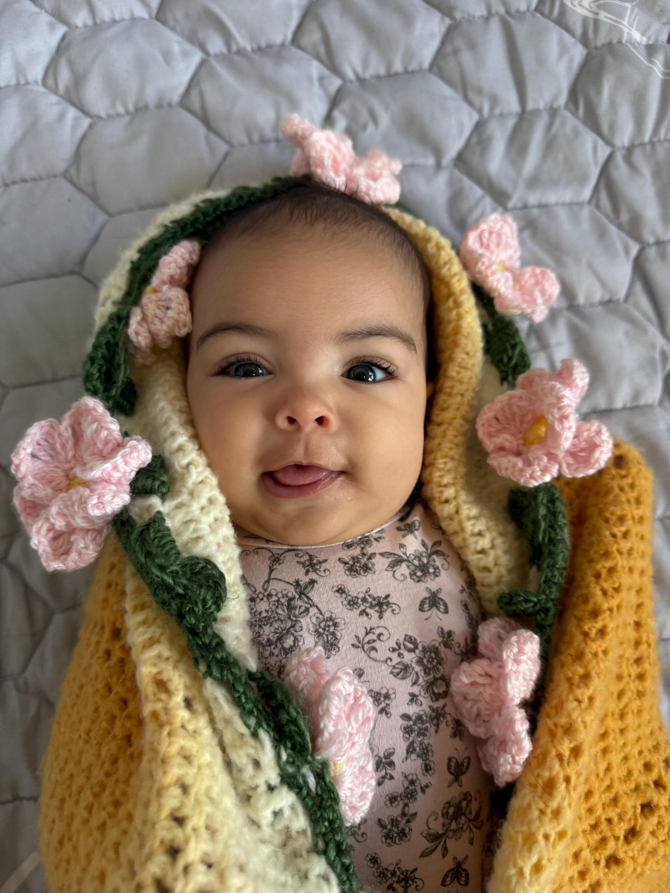 A baby lies on a gray blanket, wrapped in a yellow, white, and green crochet blanket with pink crochet flowers.