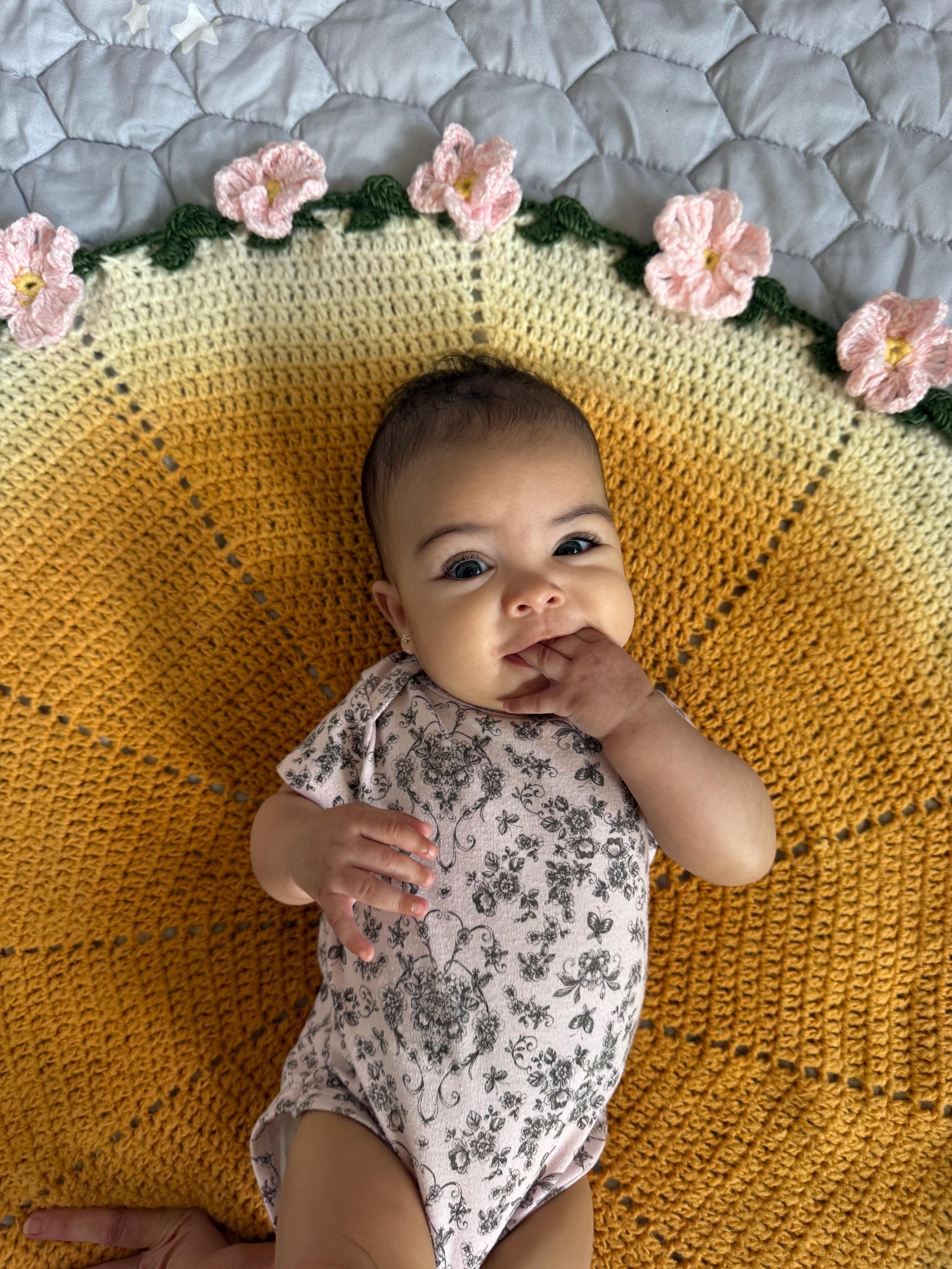 A baby in a floral onesie lies on a round, yellow and white crochet blanket with pink crochet flowers around the edge.