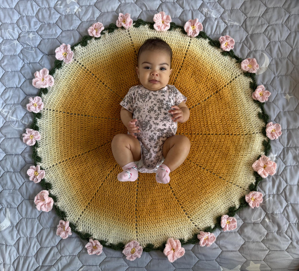 A baby in a floral onesie sits on a round, yellow and white crochet blanket with pink crochet flowers around the edge.