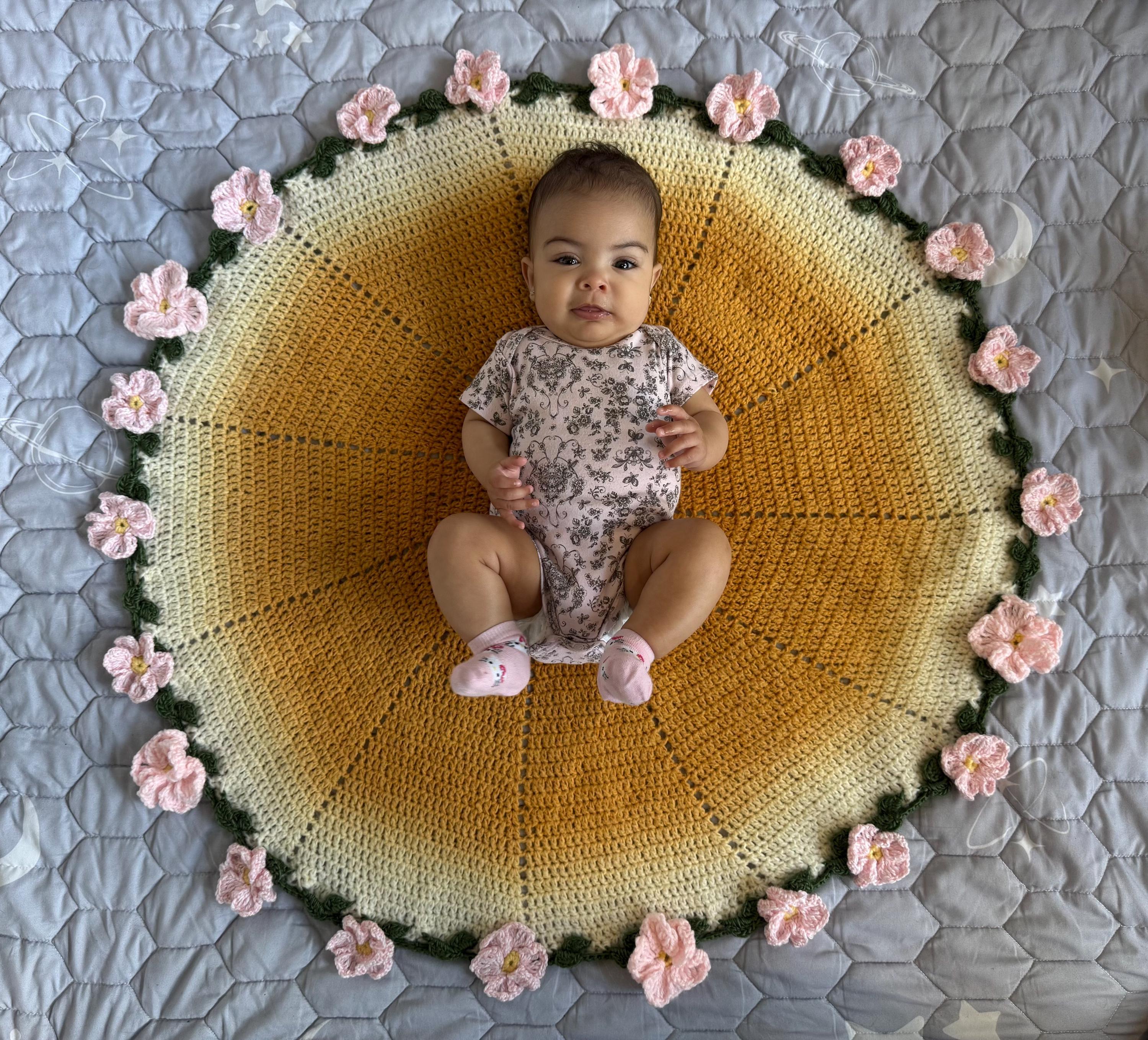 A baby in a floral onesie sits on a round, yellow and white crochet blanket with pink crochet flowers around the edge.