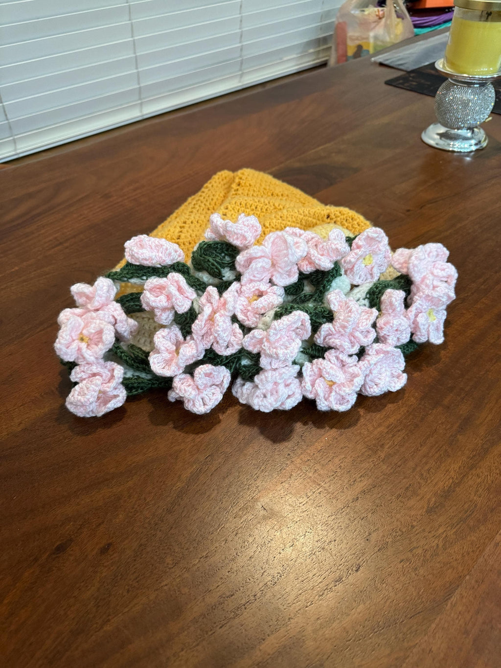 A folded yellow and white crochet blanket with a cluster of pink crochet flowers and green leaves on top, on a wooden table.