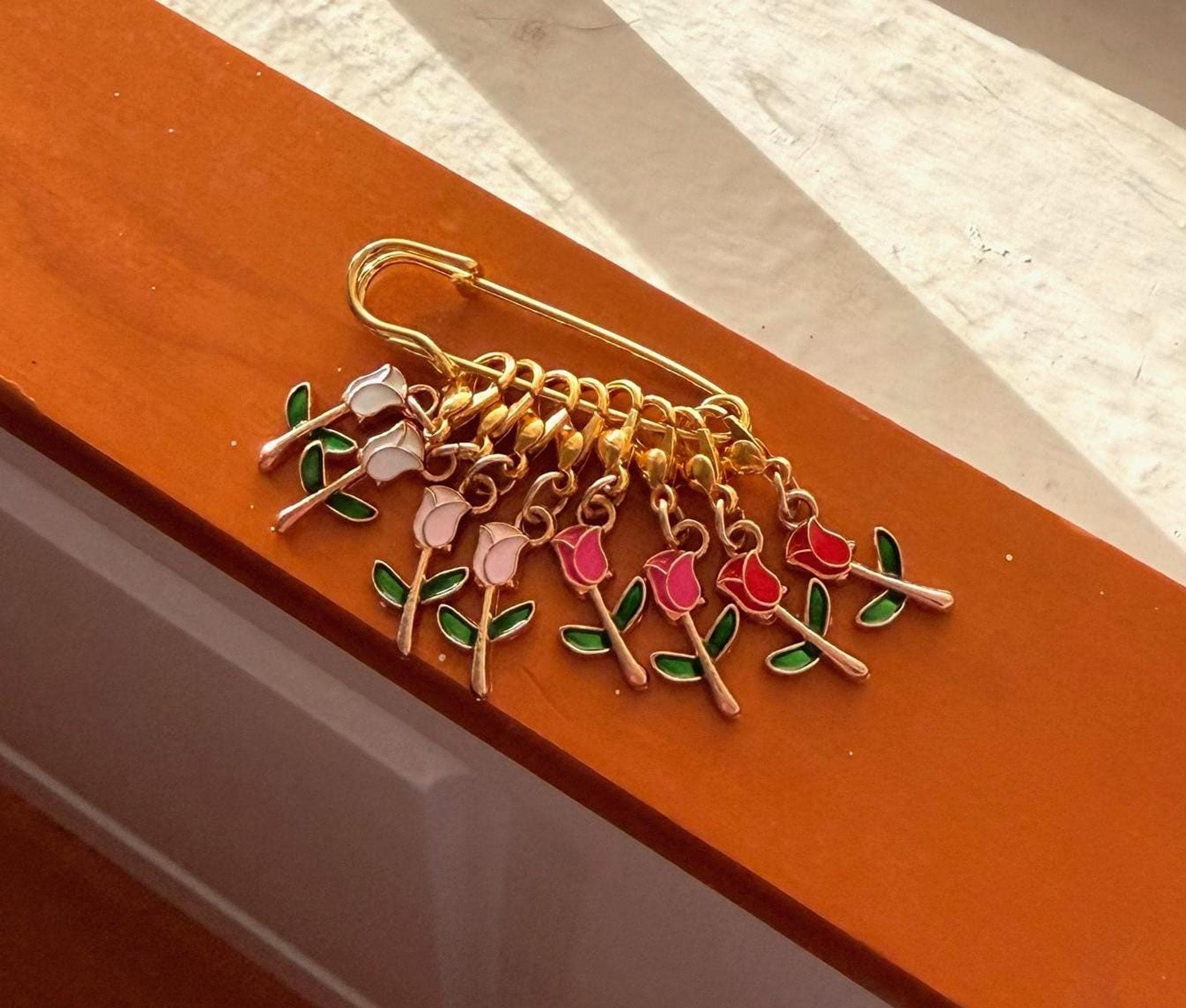 Eight enamel tulip stitch markers in shades of white, pink, and red, attached to a gold safety pin, resting on a brown surface.
