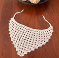 A cream-colored, crocheted, open-weave bandana with tie strings is displayed flat on a dark wooden surface.