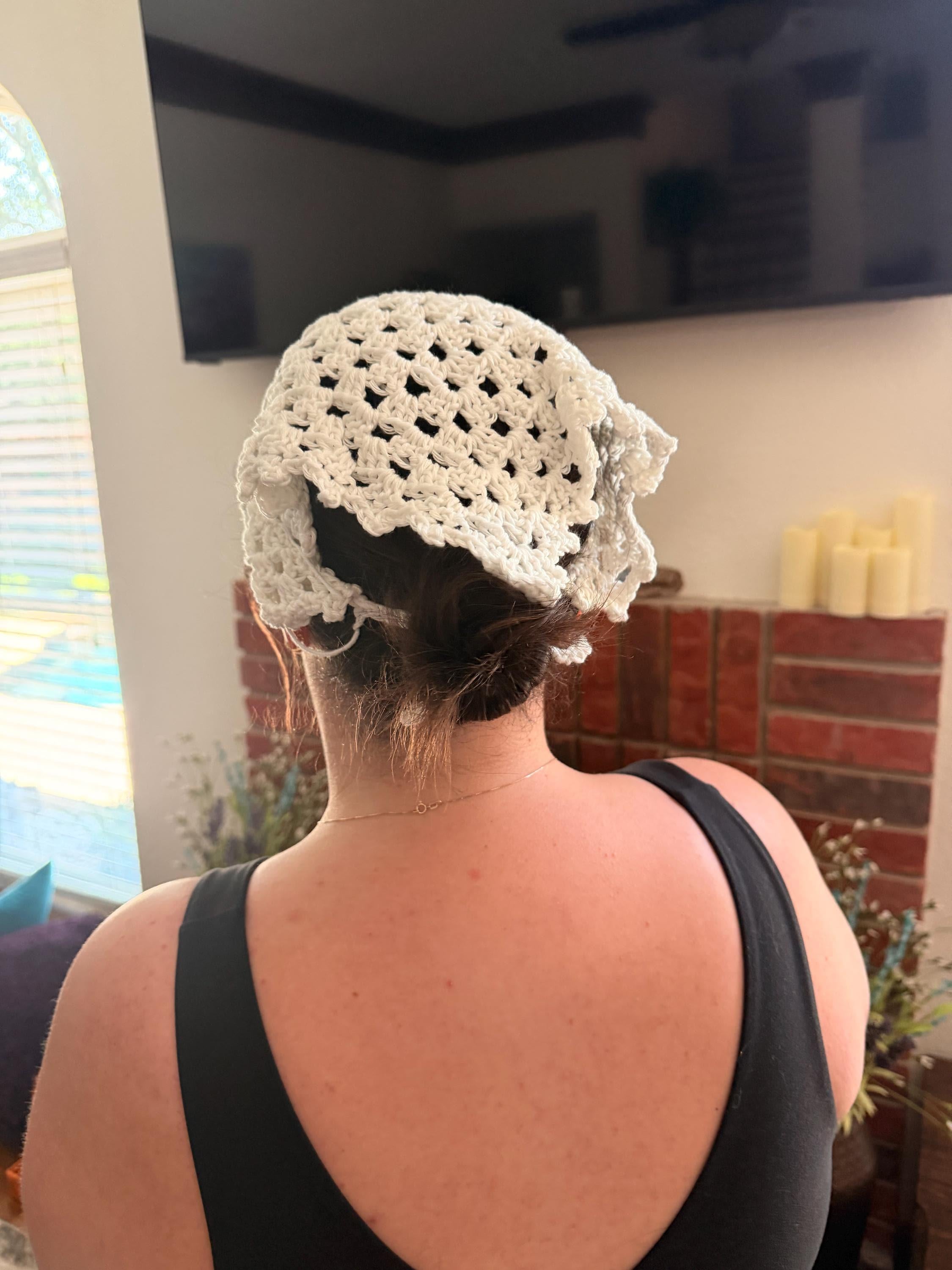 The back of a person's head and shoulders, with a cream-colored crocheted bandana tied around their head, covering their hair.