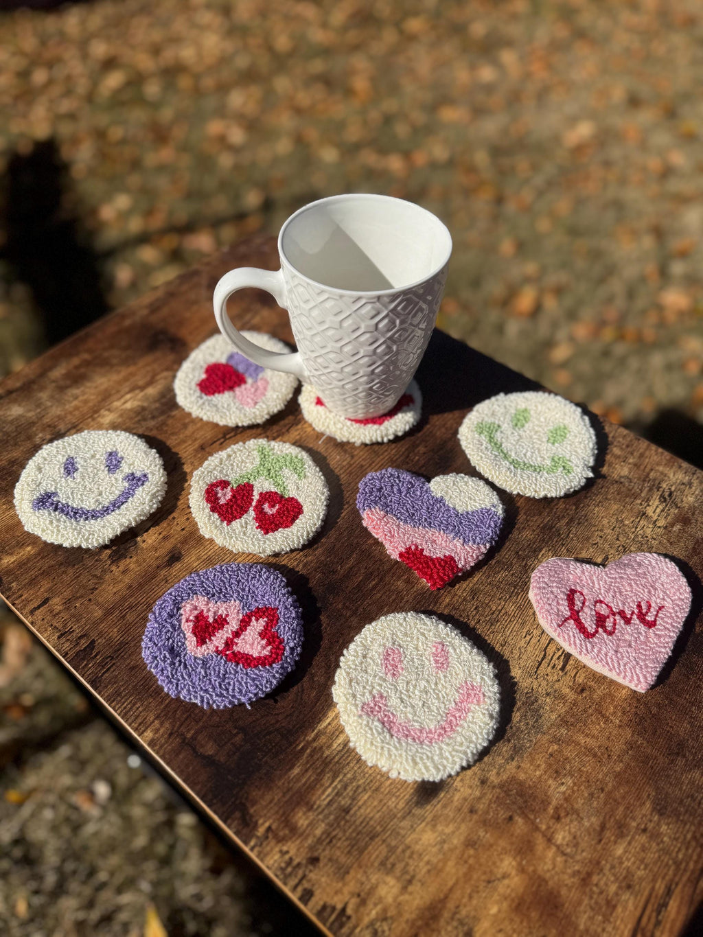 Nine punch needle coasters, including smiley faces, hearts, and cherries, are arranged on a wooden surface around a white mug.