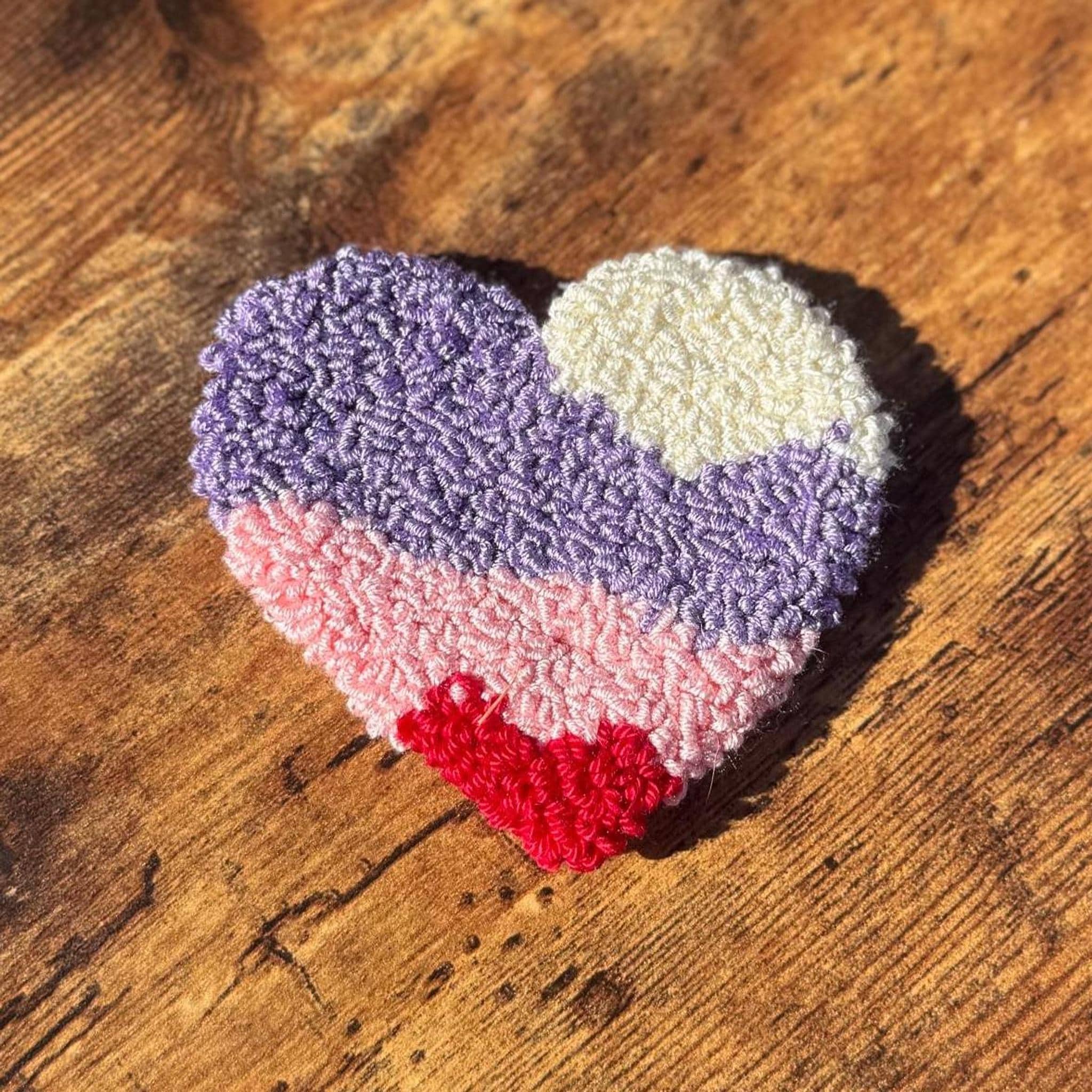 A heart-shaped punch needle coaster with horizontal stripes of purple, pink, and red on a wooden surface.