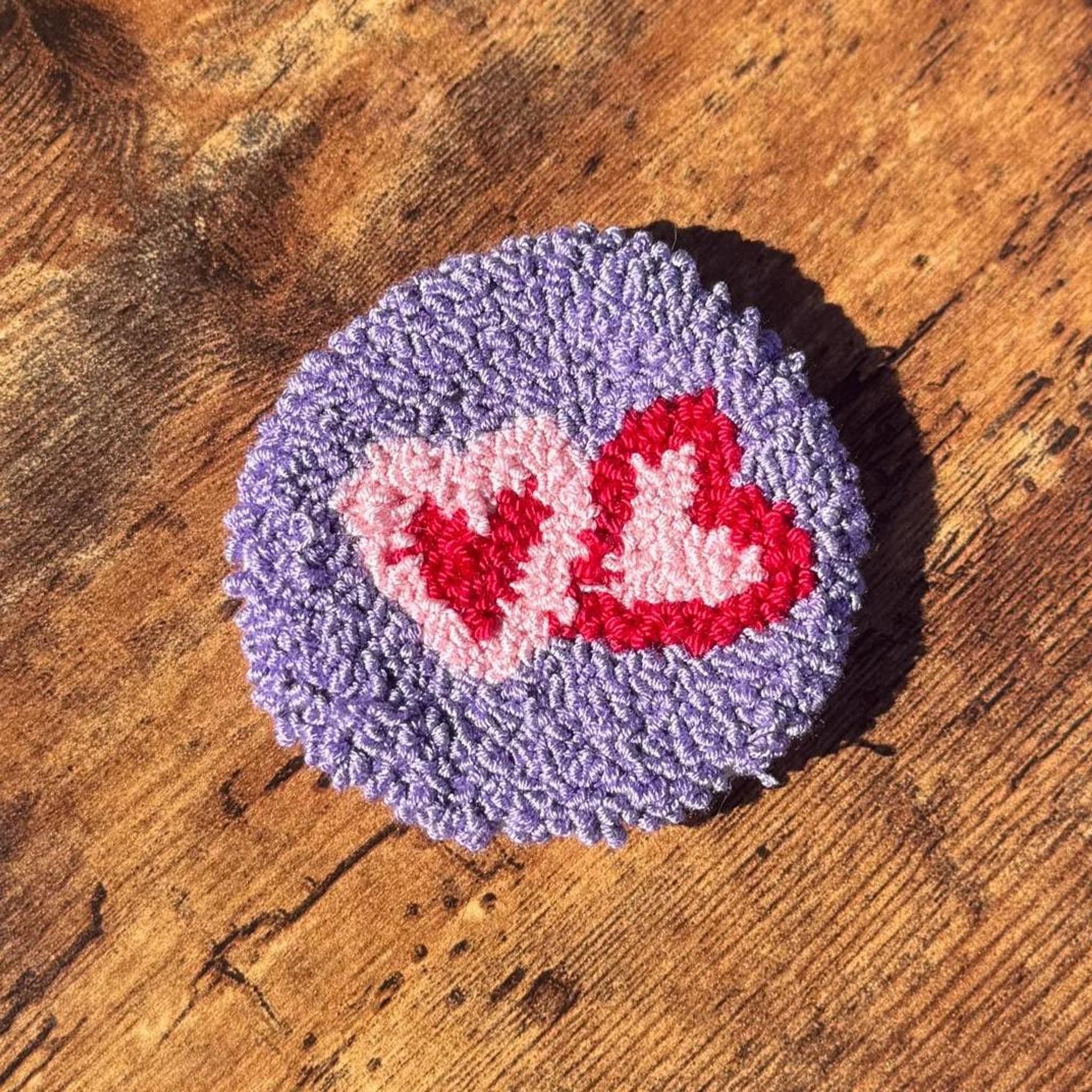 A round purple punch needle coaster with two pink hearts outlined in red on a wooden surface.