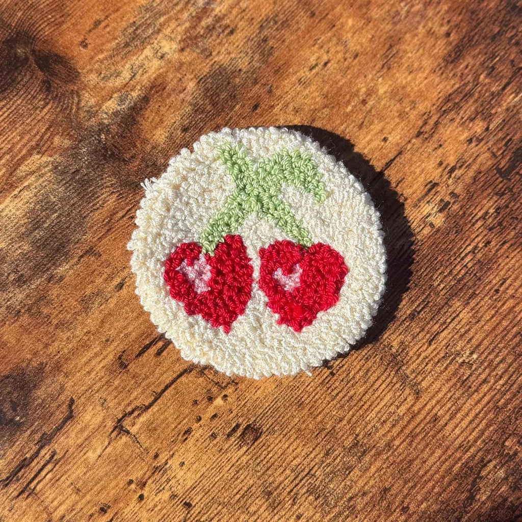 A round white punch needle coaster with two red cherries and green stems on a wooden surface.