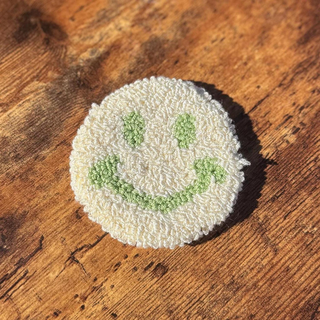 A round white punch needle coaster with a green smiley face on a wooden surface.
