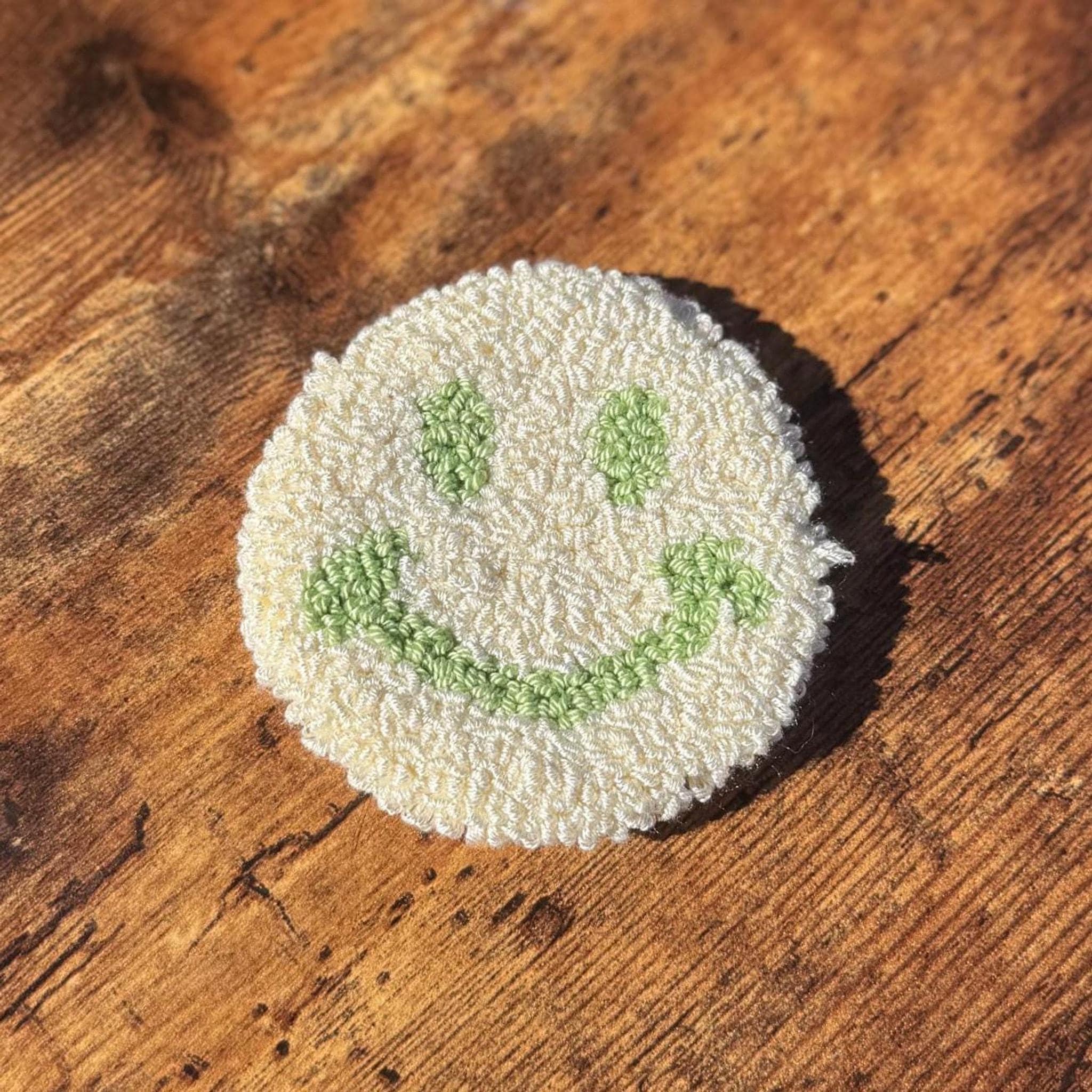 A round white punch needle coaster with a green smiley face on a wooden surface.