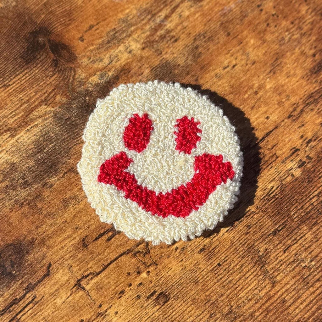 A round white punch needle coaster with a red smiley face on a wooden surface.