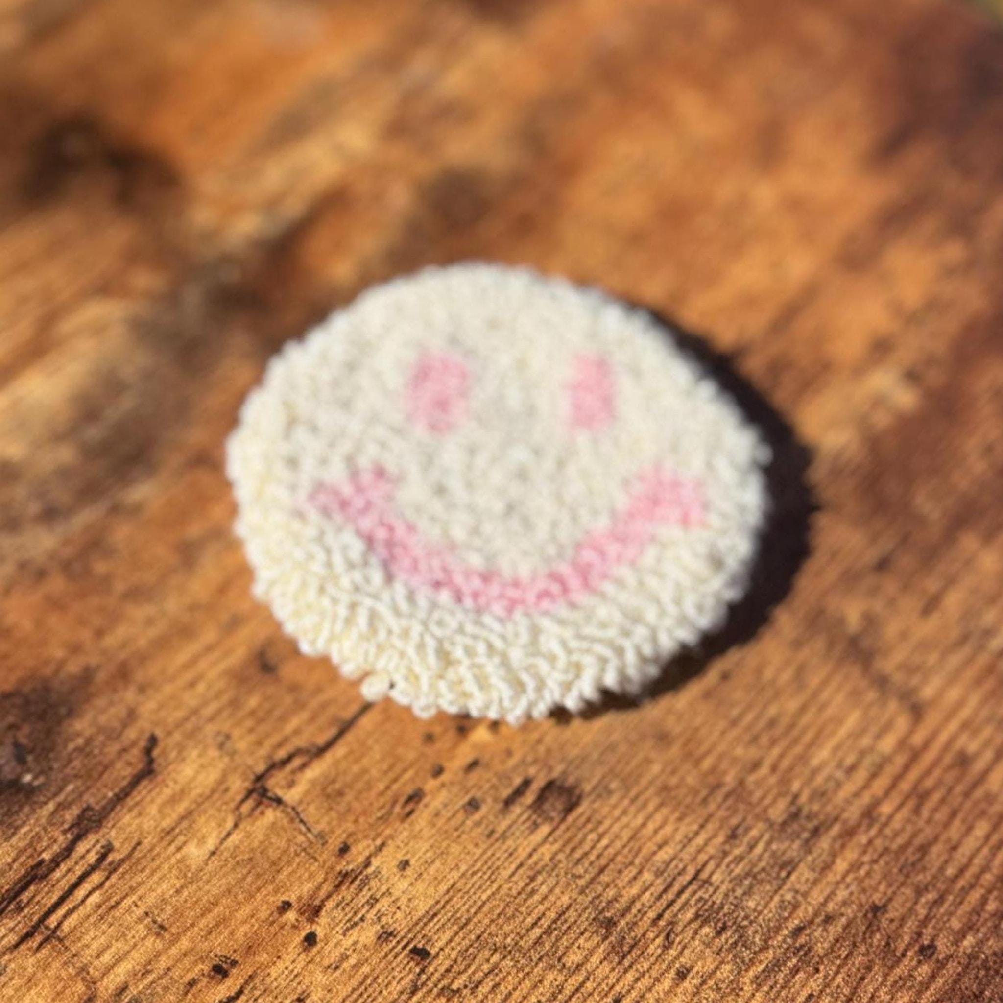 A round white punch needle coaster with a pink smiley face, slightly out of focus, on a wooden surface.