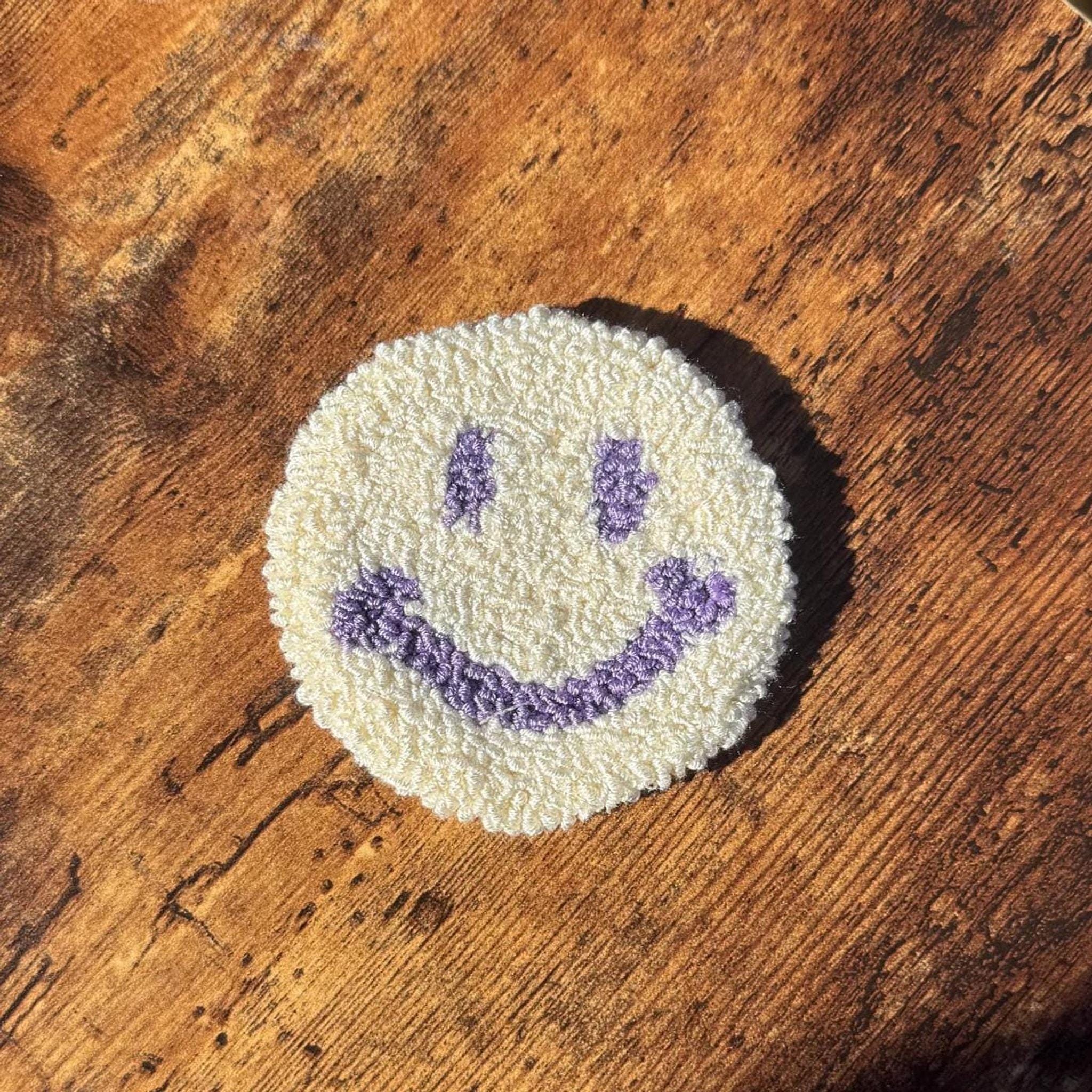 A round white punch needle coaster with a purple smiley face on a wooden surface.