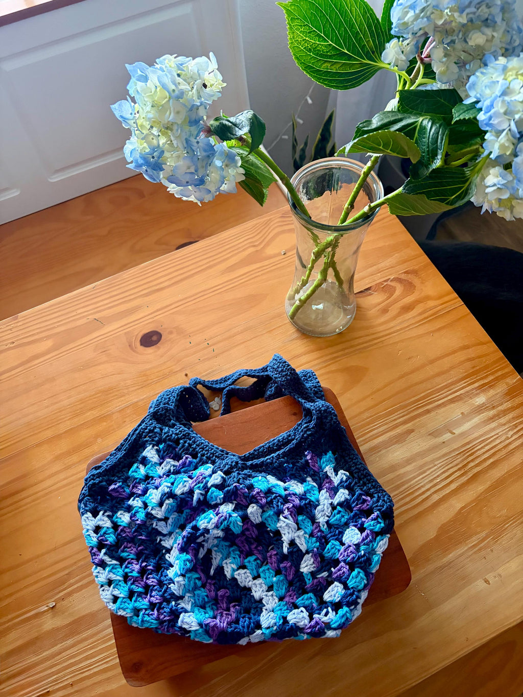 A dark blue crochet market bag with light blue, purple, and white accents sits on a wooden table next to a vase of blue hydrangeas.