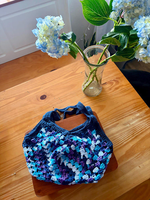 A dark blue crochet market bag with light blue, purple, and white accents sits on a wooden table next to a vase of blue hydrangeas.