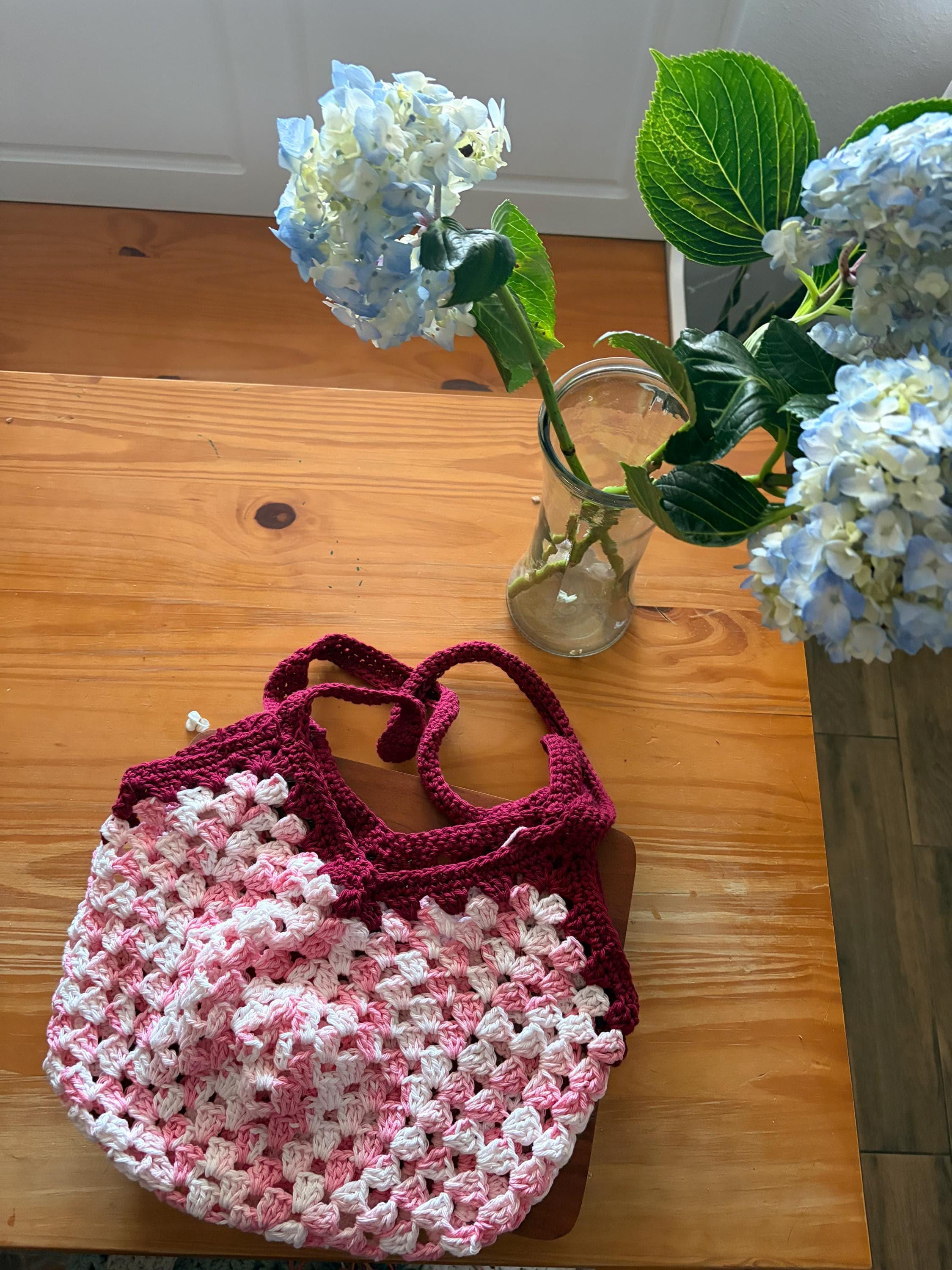 A dark pink crochet market bag with light pink and white accents sits on a wooden table next to a vase of blue hydrangeas.