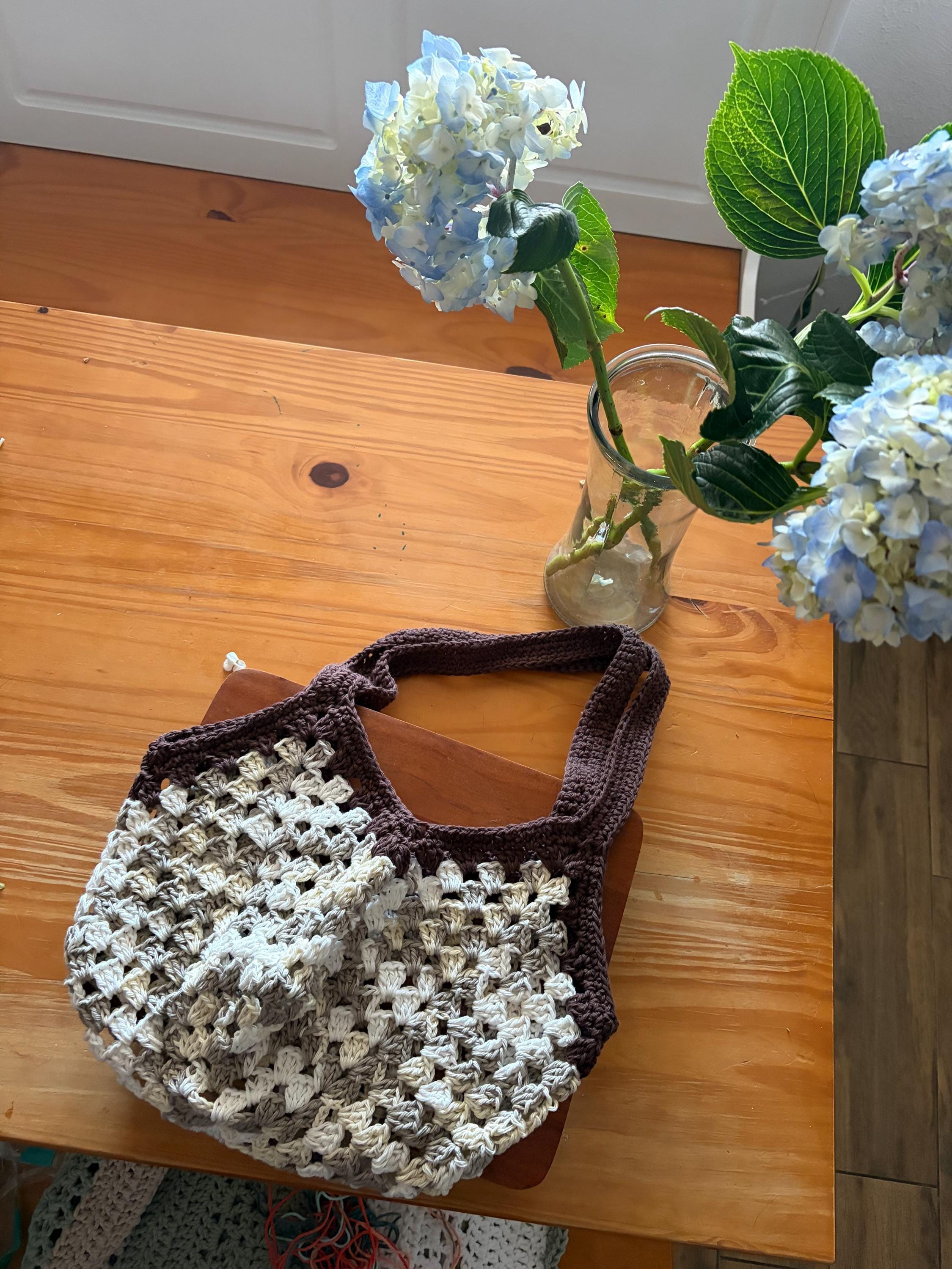 A brown crochet market bag with white and tan accents sits on a wooden table next to a vase of blue hydrangeas.
