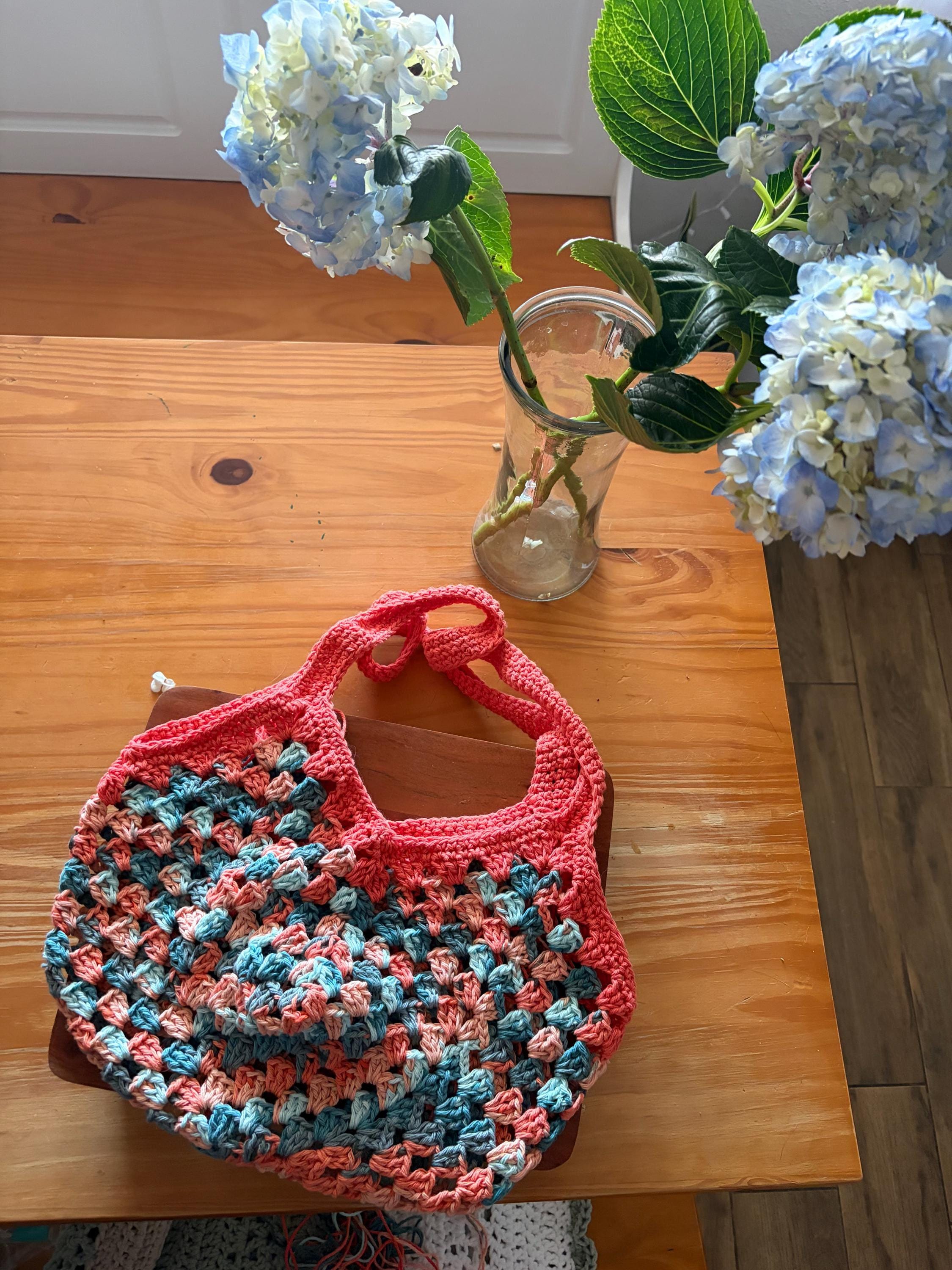 A coral crochet market bag with light blue and tan accents sits on a wooden table next to a vase of blue hydrangeas.