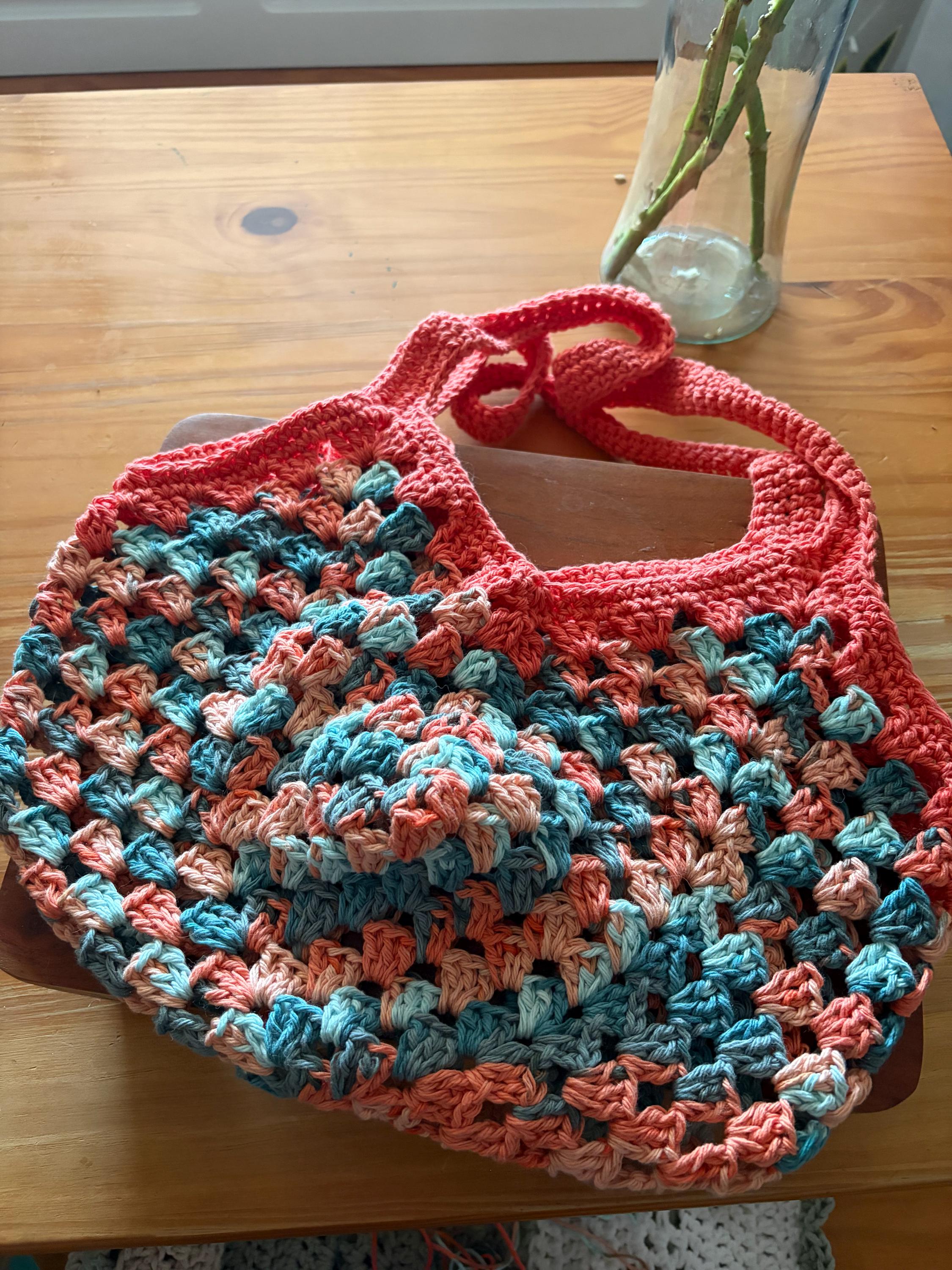 A coral crochet market bag with light blue and tan accents is centered on a wooden table.
